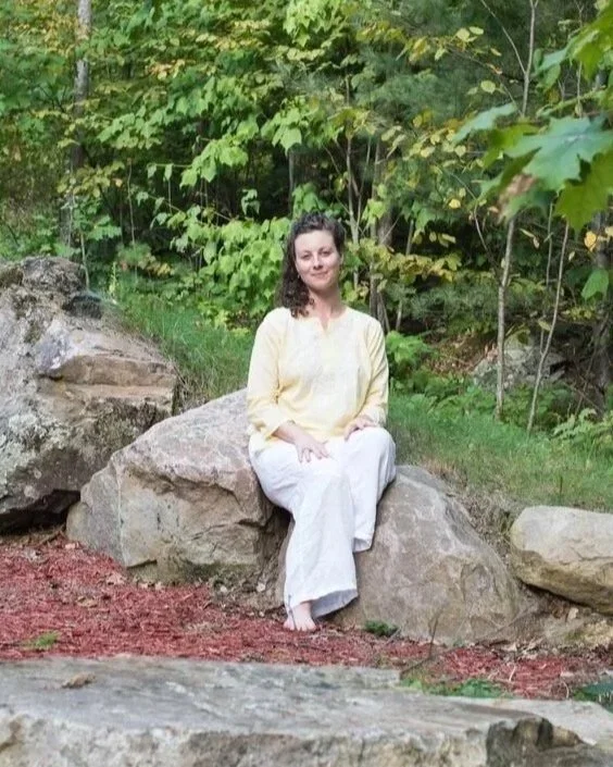 A woman wearing a yellow top and white pants sitting on a large rock in a wooded area with green foliage and trees around her.
