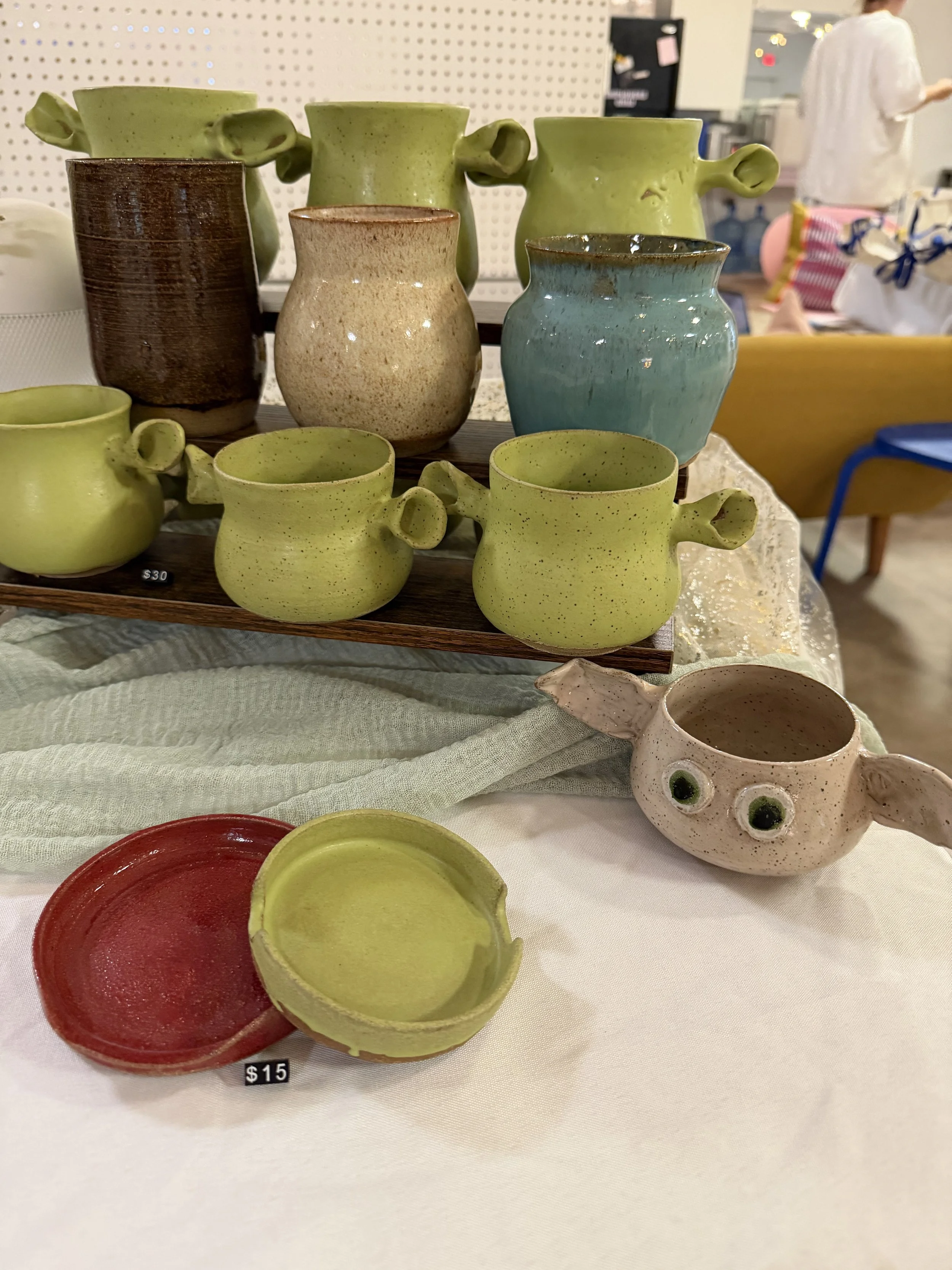 Handmade ceramic cups in various colors and shapes displayed on a table at an art fair.