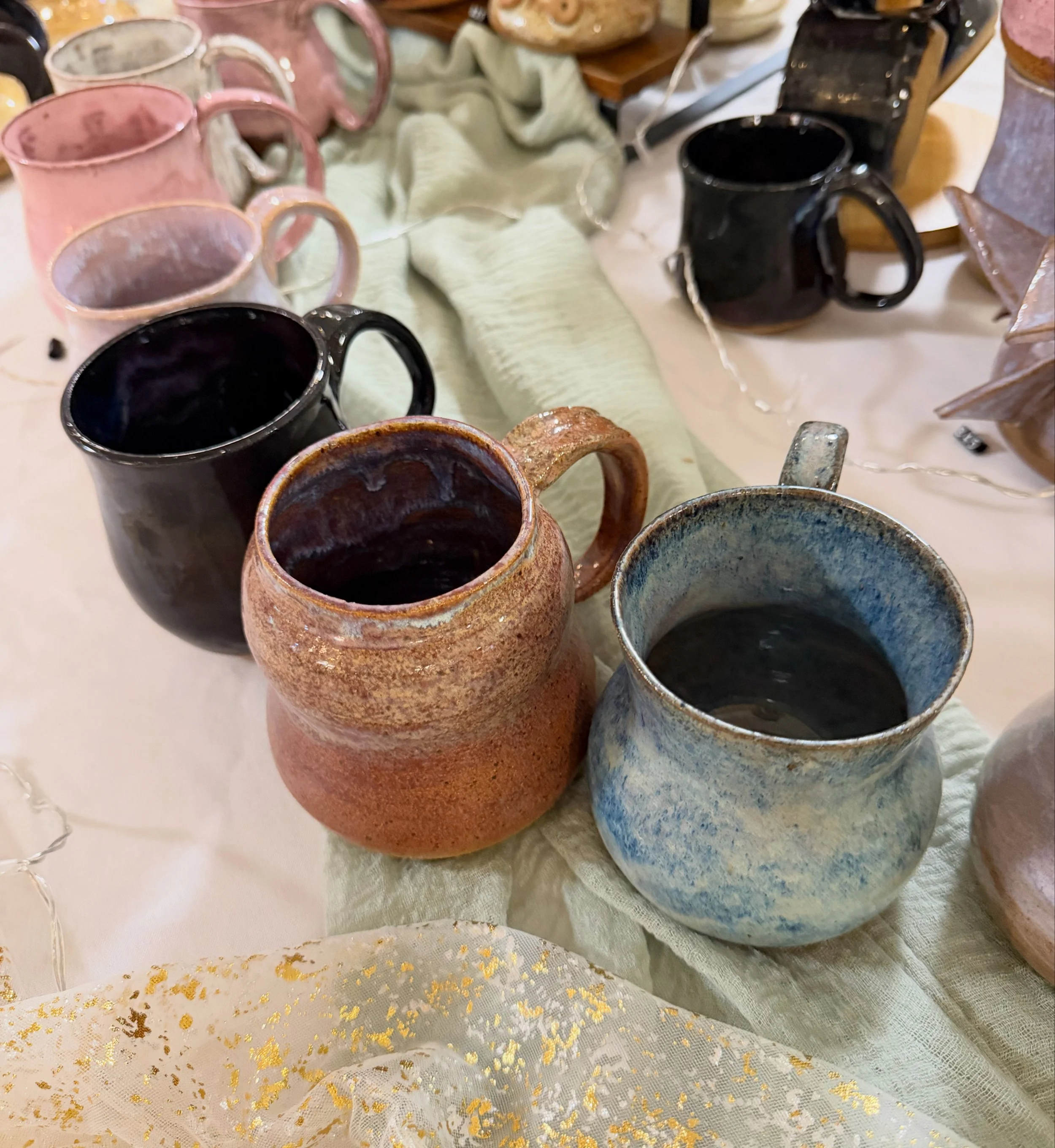 Collection of ceramic mugs in various colors and styles displayed on a table with a cloth and decorative gold accents.