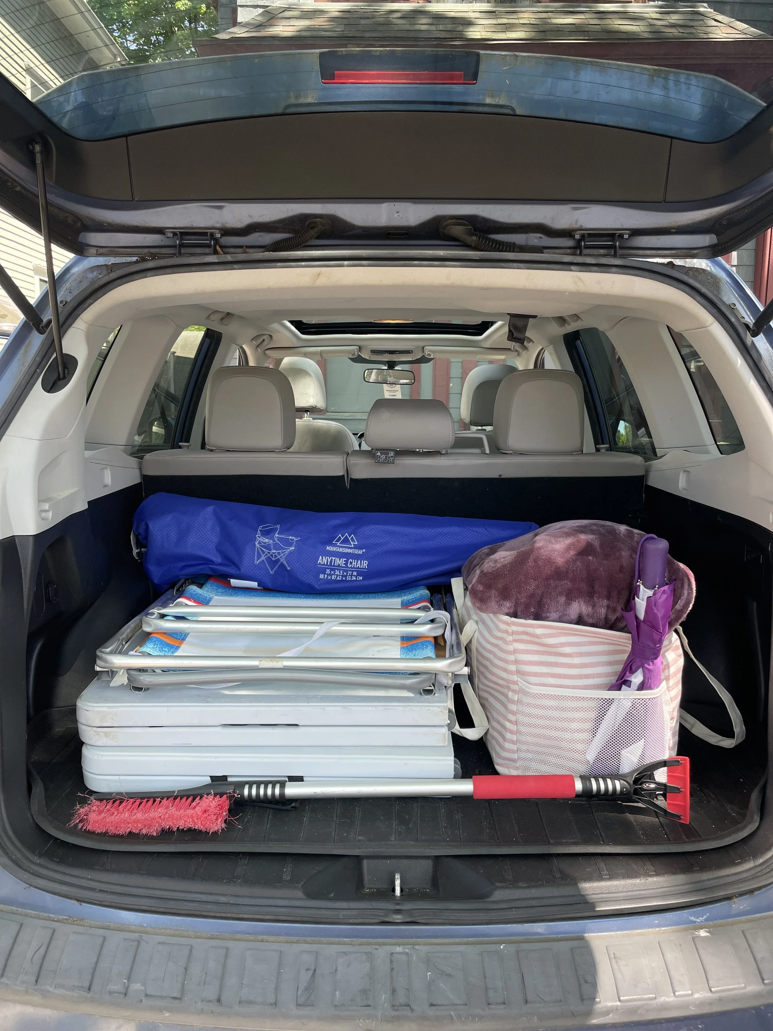 The open trunk of a car filled with various items including a blue camping tent, white coolers, a pink and white striped bag with a fuzzy pink cover, and a red snow shovel. The interior of the car shows four beige leather seats and a rearview mirror.