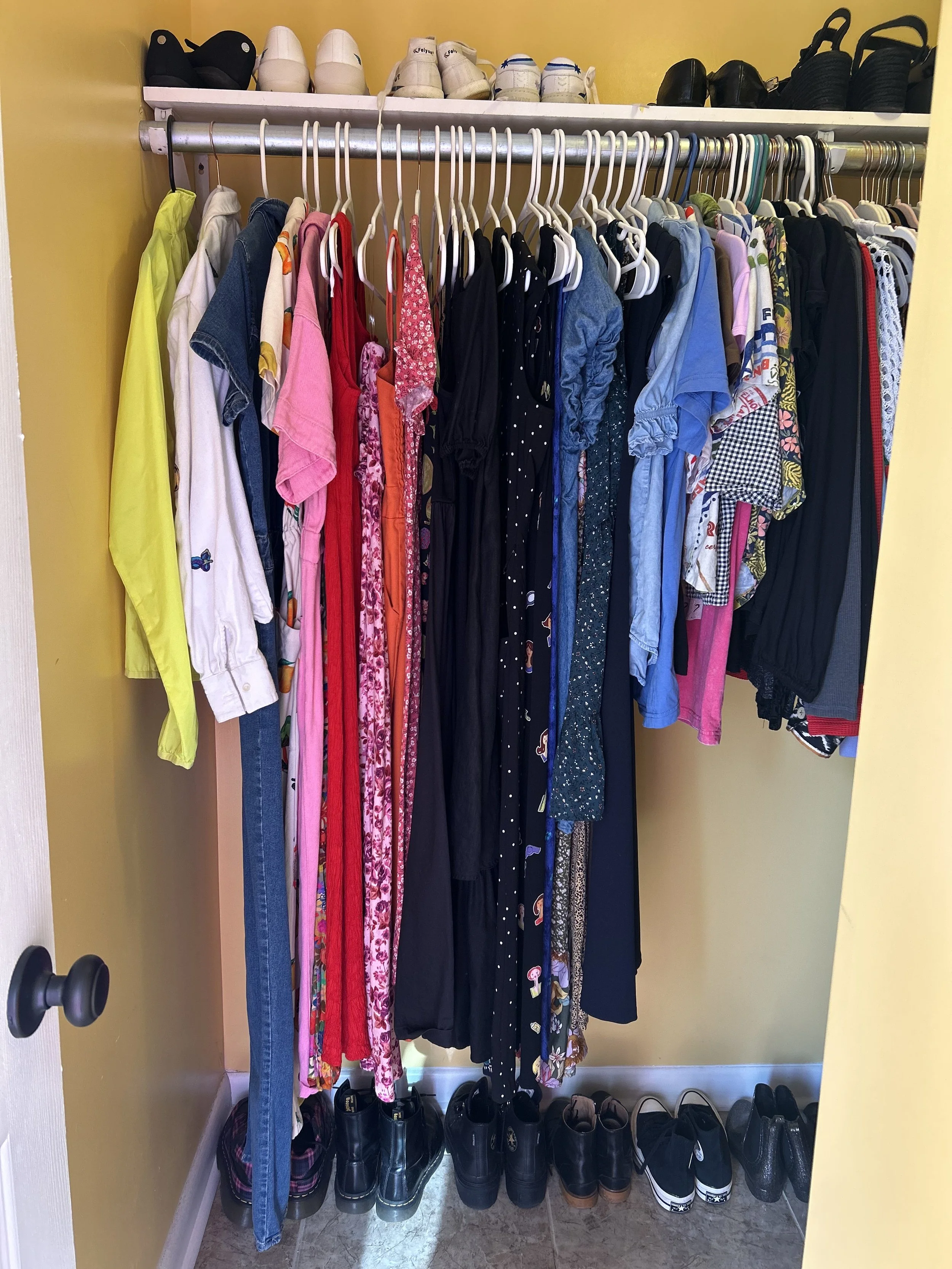 A closet with a yellow wall, containing a shelf with shoes on top, a horizontal rod with hanging clothes including various colors and styles, and shoes neatly arranged on the floor below.