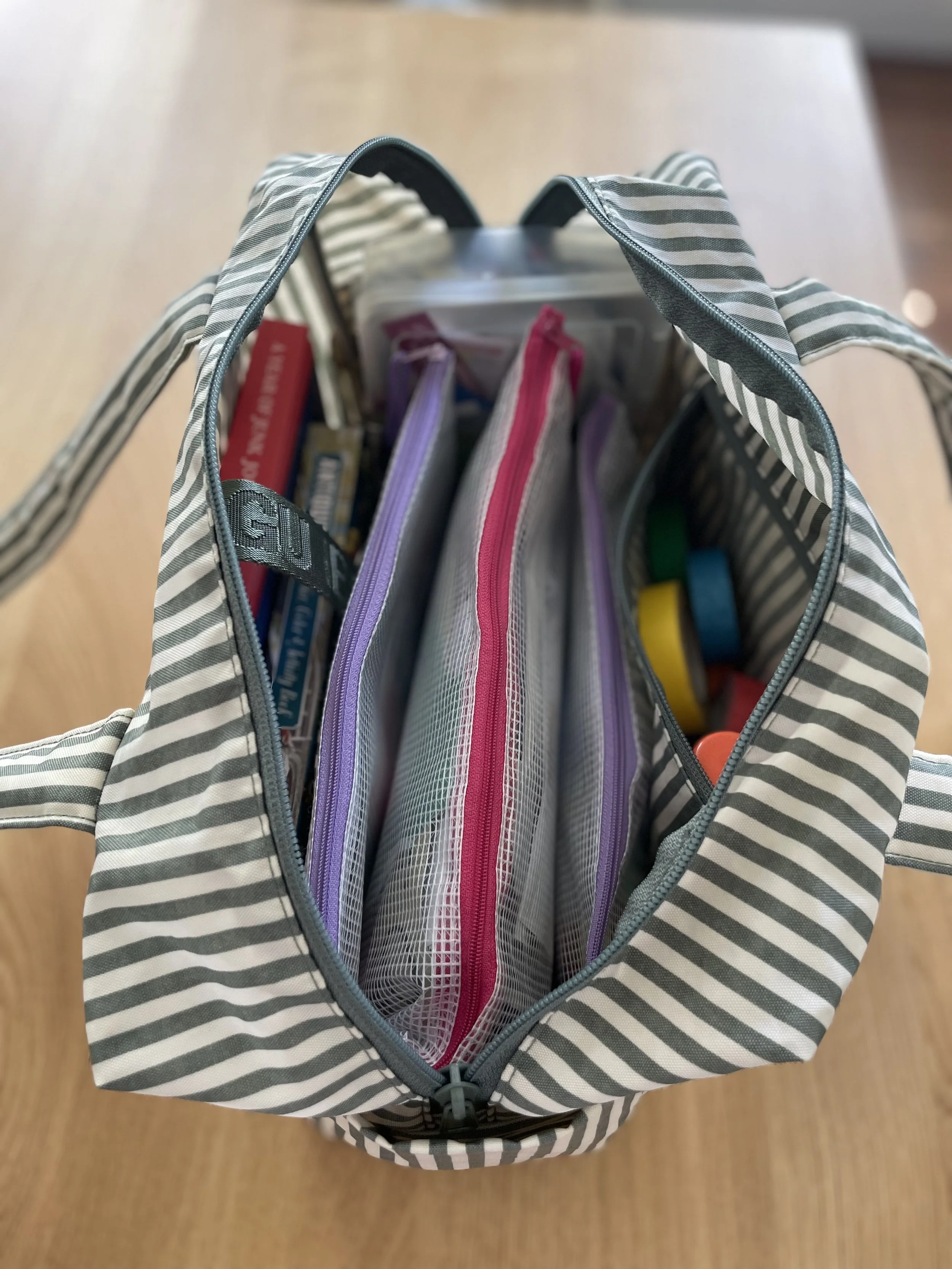 A striped gray and white tote bag, open at the top, with various school supplies inside including notebooks, folders, and a pair of scissors.