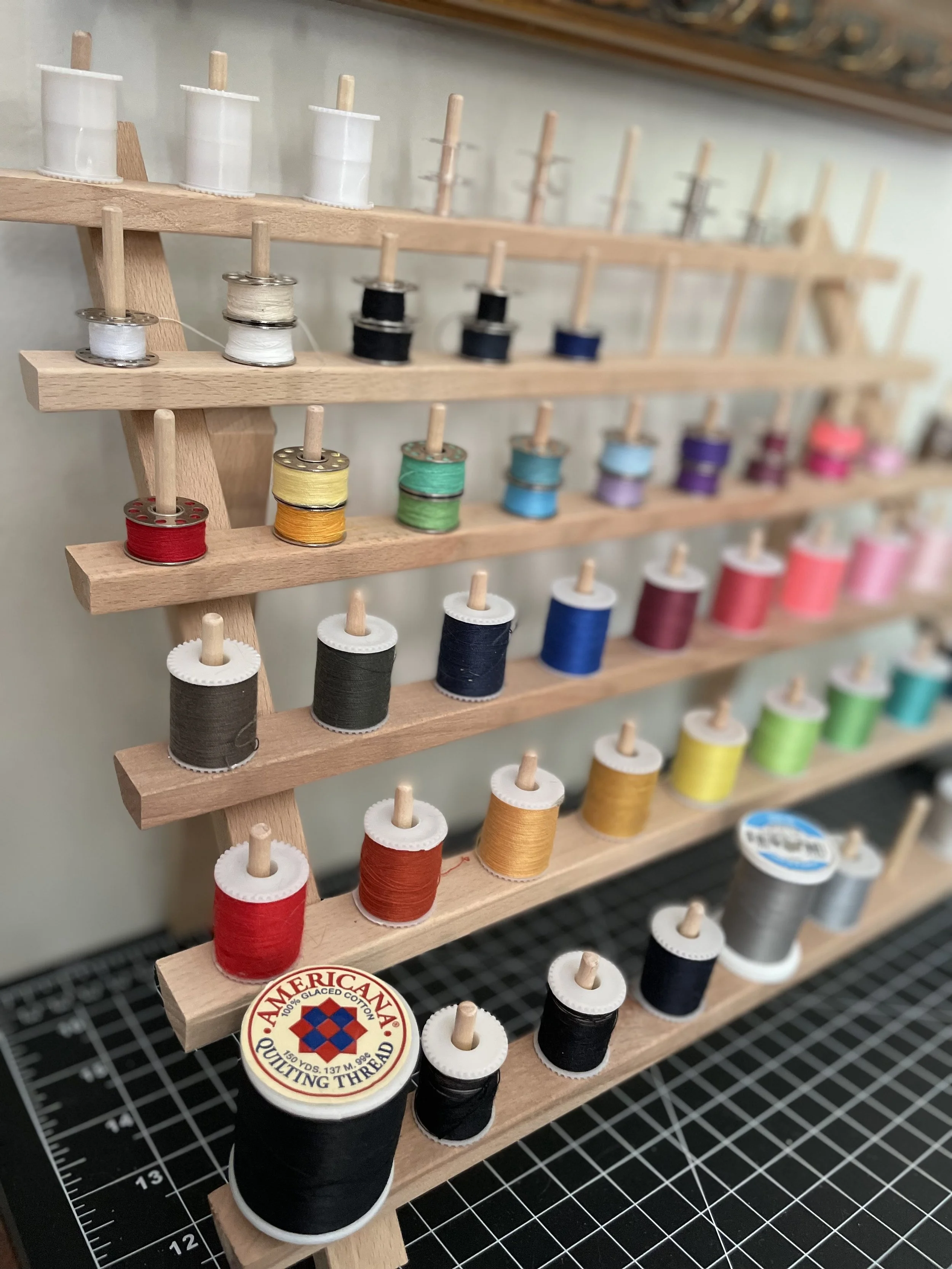 A wooden thread rack holding spools of various colored sewing threads arranged in rows from light to dark colors, with some spools labeled and a cutting thread tin at the bottom.