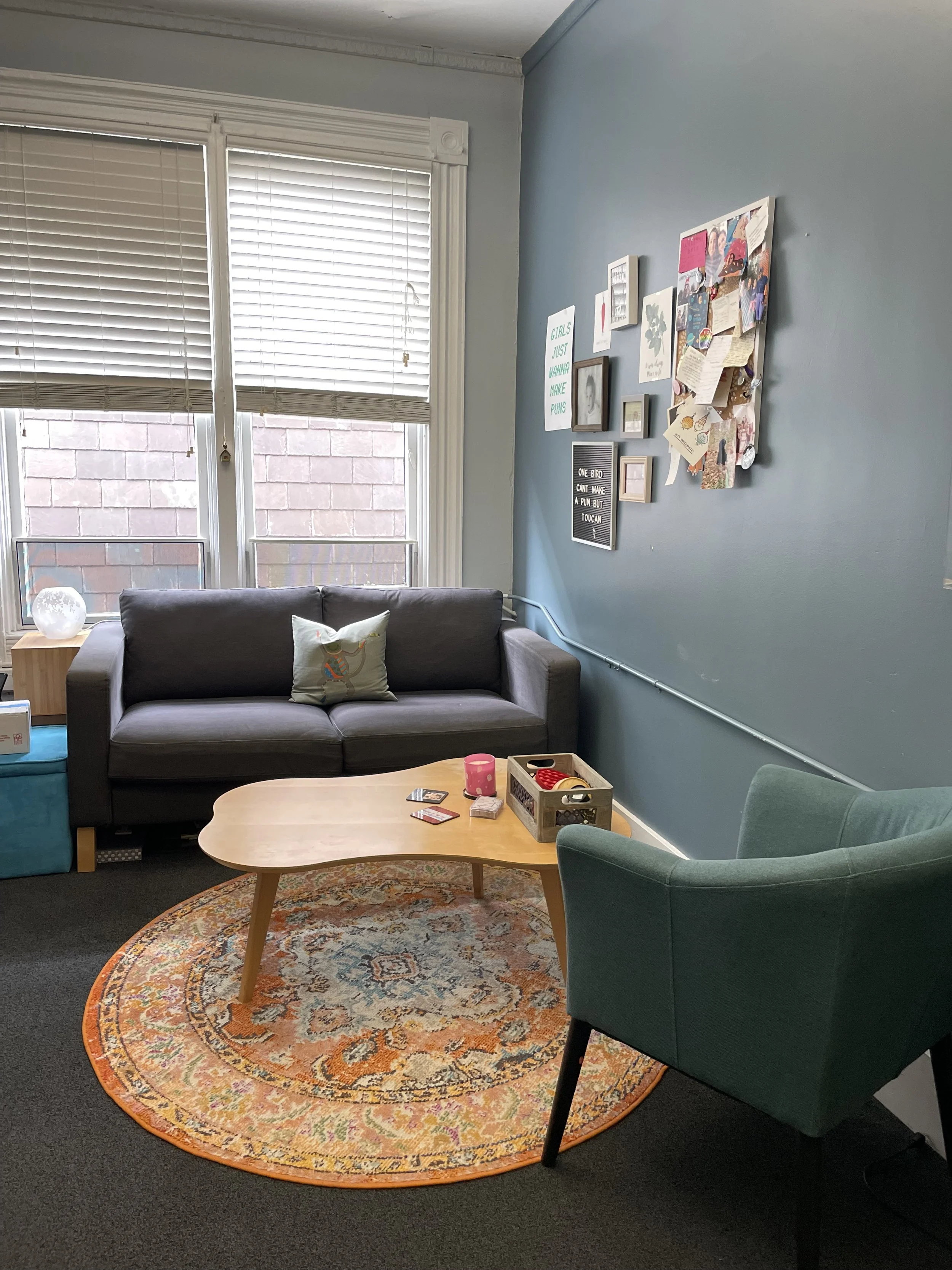 Living room with a gray couch, a green armchair, a round orange and multicolored rug, a light wood coffee table with toys and cards, a wall with various pictures and notes, windows with closed blinds, and a lamp on a side table.