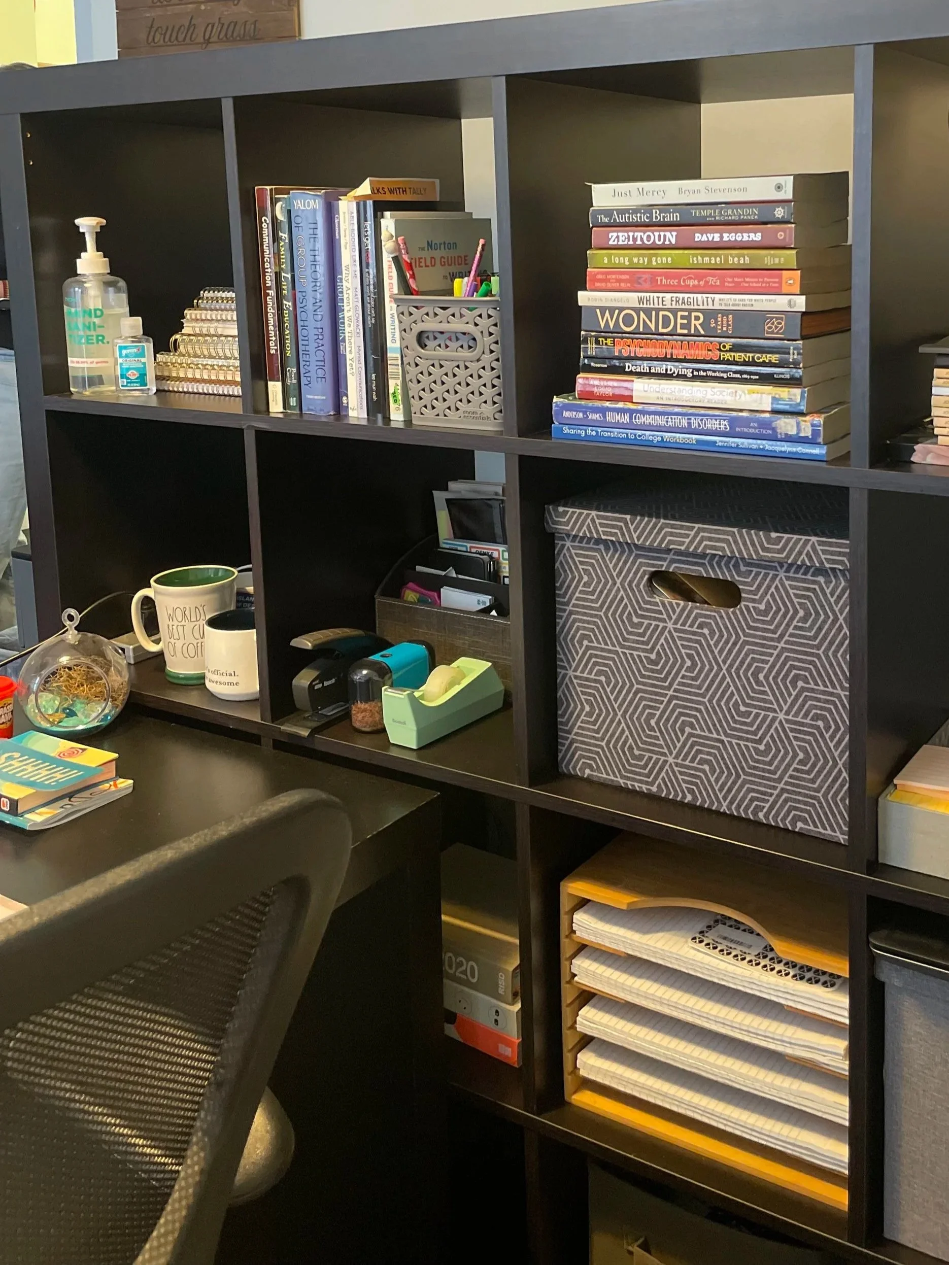 A black bookshelf with multiple compartments containing a variety of books, office supplies, and organizational boxes. A black desk with a chair is in front of the bookshelf, with notebooks and office items on it.