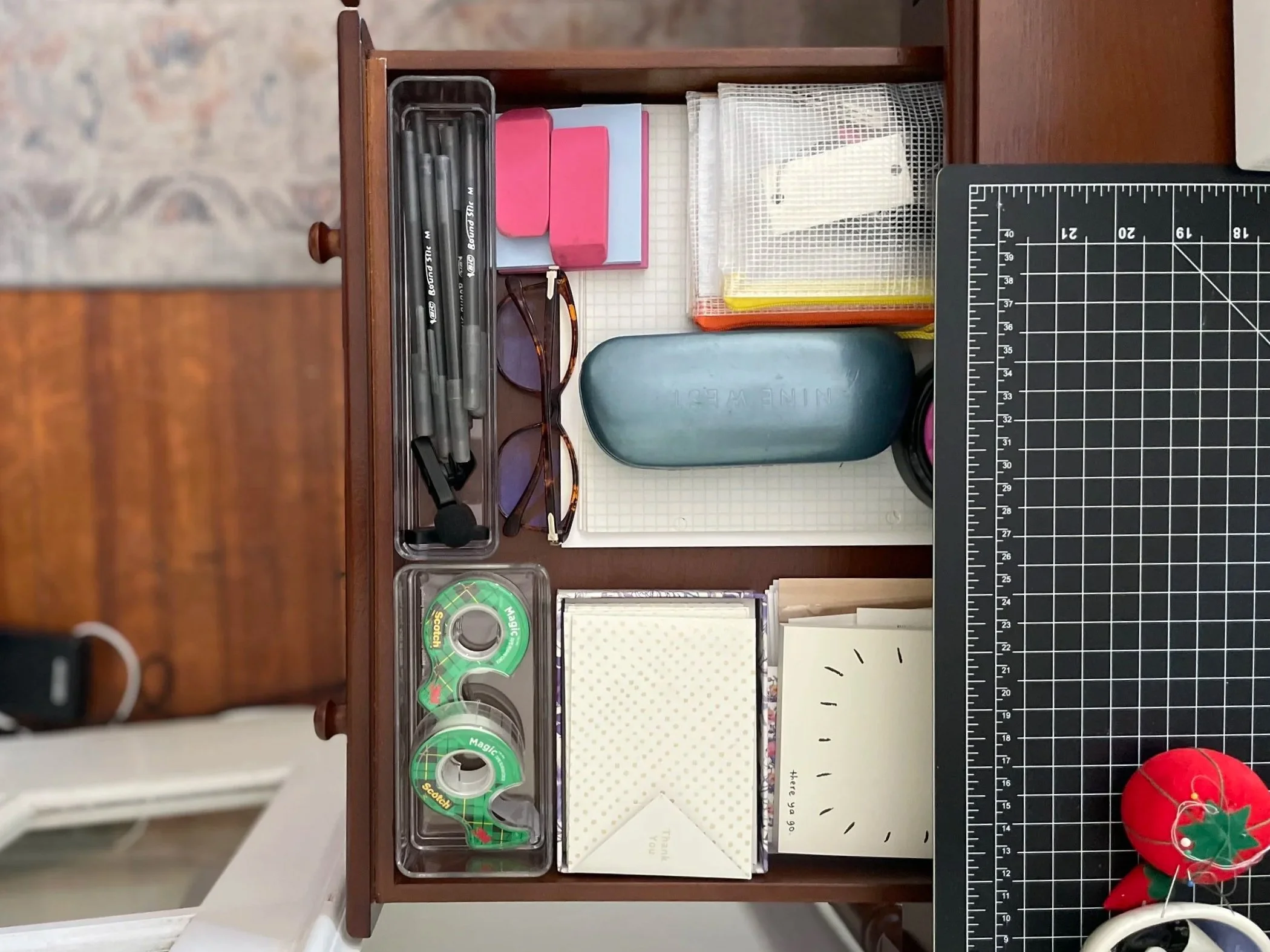 A wooden desk drawer containing various office supplies including multiple pens, a pair of eyeglasses, pink and white boxes, a mesh pencil holder, tape, note cards, and a cutting mat with measurement markings.