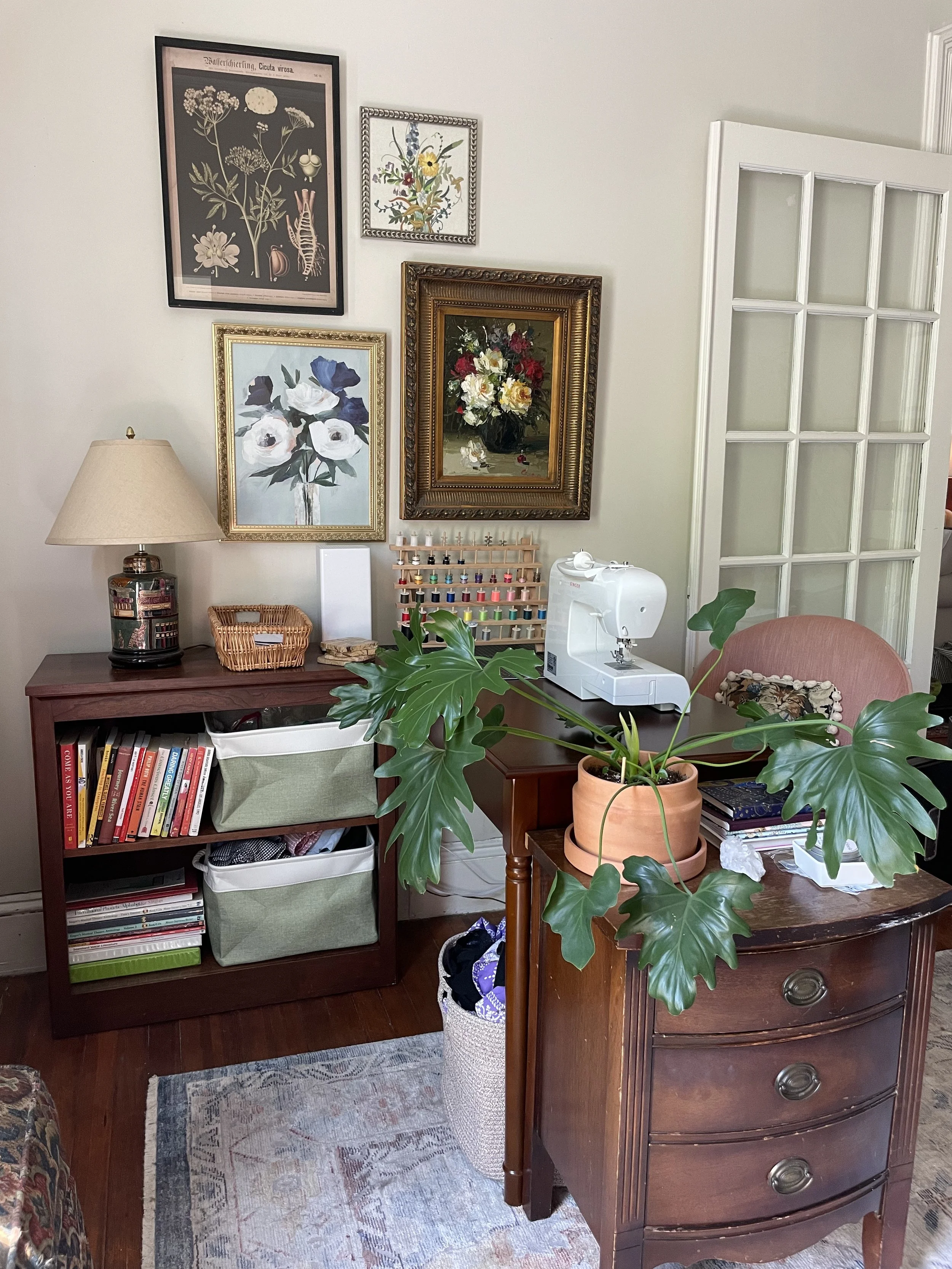 A corner of a room with a wooden dresser, green houseplant, lamp, and a bookshelf. The wall has framed botanical artwork.