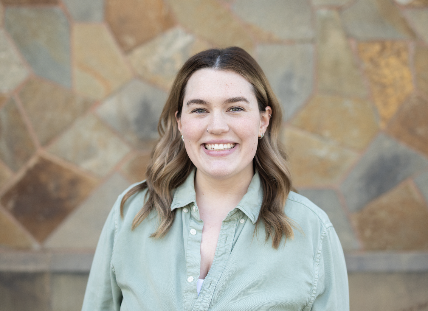 A smiling woman with brown wavy hair, wearing a light green button-up shirt, standing in front of a stone wall.