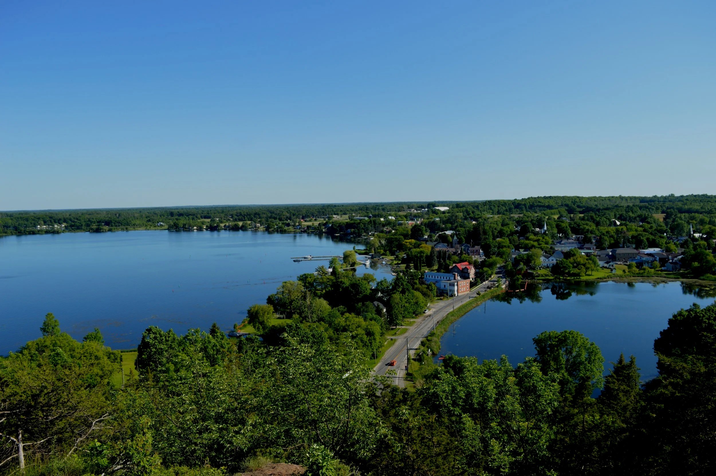 A scenic aerial view of a town beside a large body of water, with a clear blue sky and lush green trees.