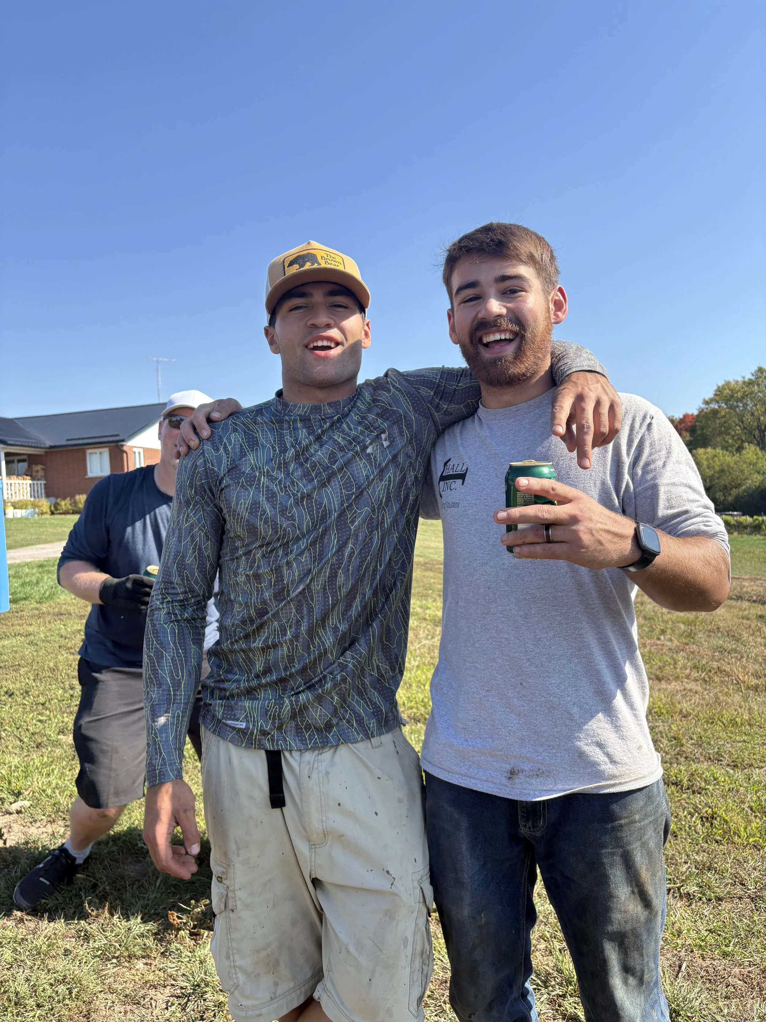 Two young men standing outdoors with their arms around each other, smiling. One is wearing a gray t-shirt, the other is wearing a patterned long-sleeve shirt and a tan cap. In the background, a third person is visible wearing sunglasses and a white cap. A house and trees are also visible under a clear blue sky.