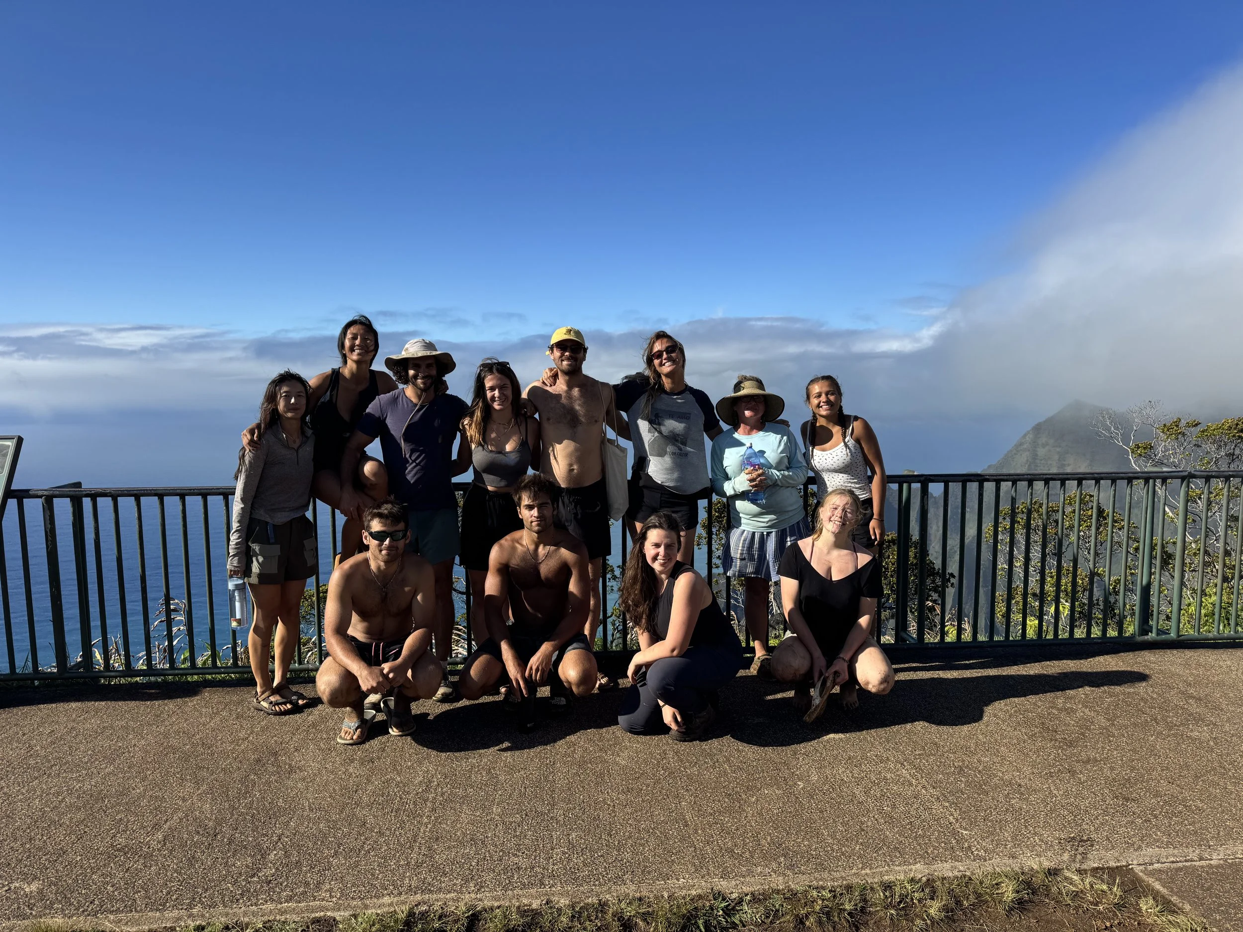 Group of eleven people posing together outdoors on a sunny day, with ocean and mountain scenery in the background, standing behind a black railing.