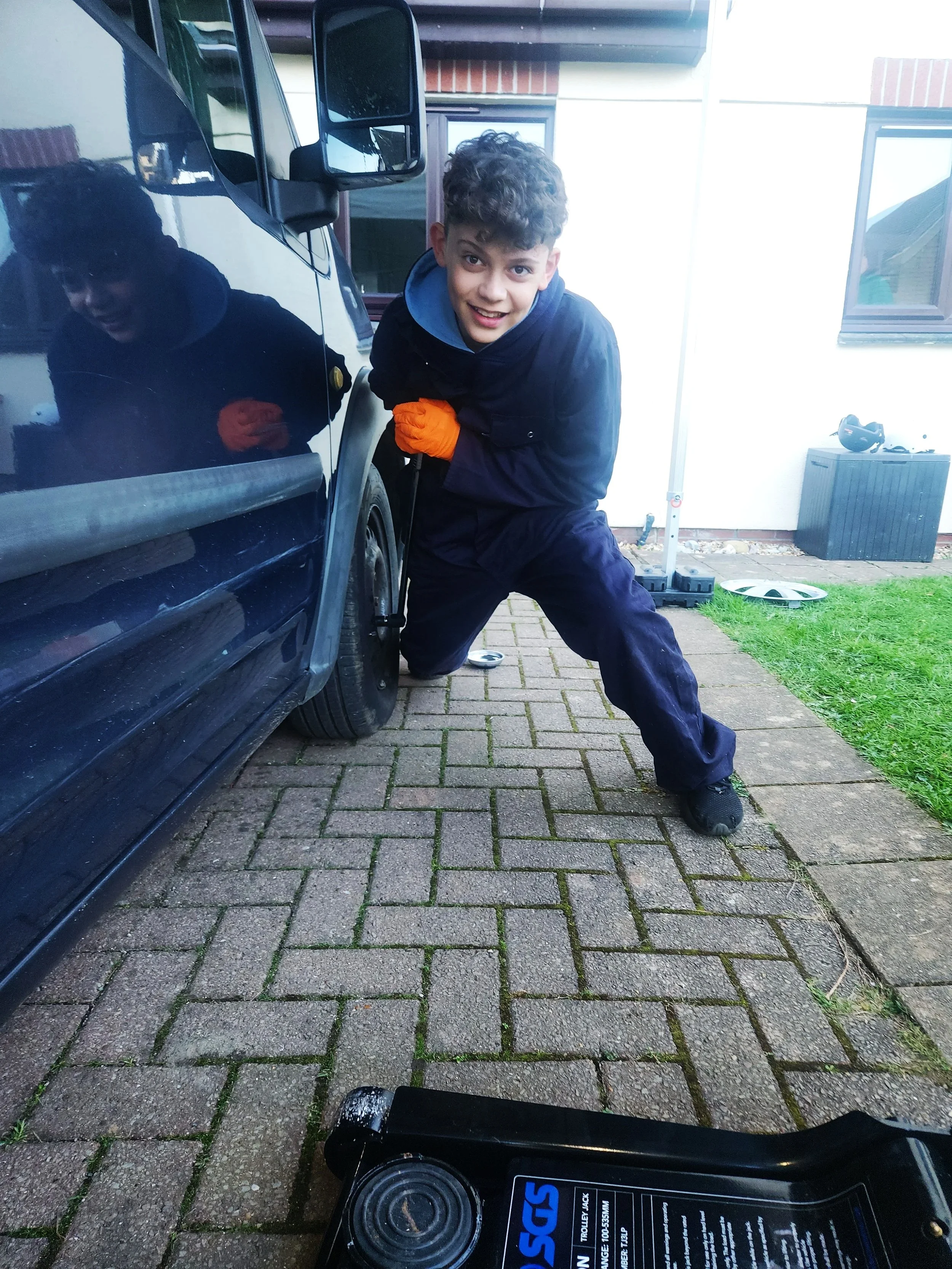 A boy wearing a dark jacket and orange gloves is kneeling next to a black vehicle, using a tool on the wheel. He is smiling at the camera. The scene is outdoors on a paved area with grass and a house in the background, and there are tools and a box on the ground nearby.
