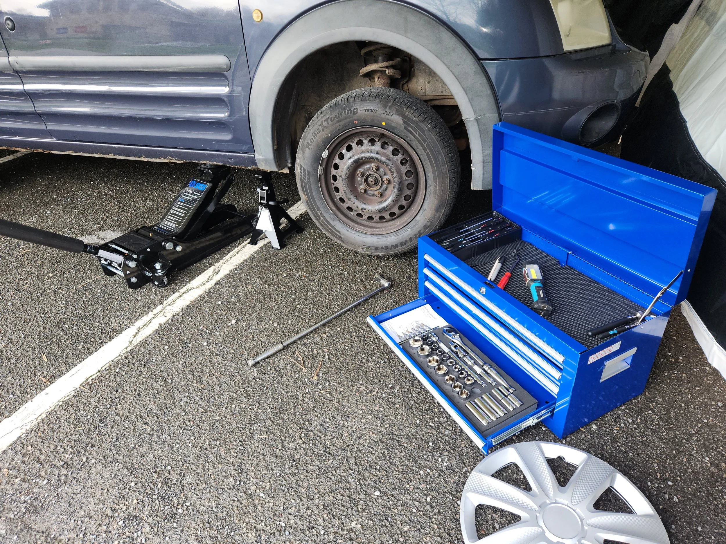 Automotive tools including a blue mechanic's toolbox, socket set, trolley jack, and axle stand next to a car wheel.