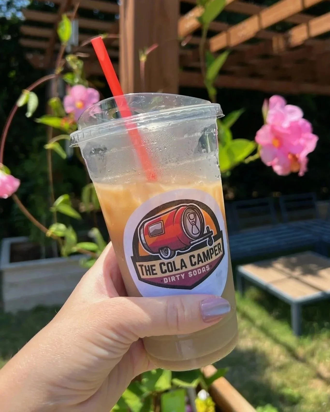 A hand holding a clear plastic cup with a red straw, containing a light orange soda. The cup has a label with a logo of a soda can and the text "The Cola Camper" and "Dirty Sodas." In the background, there are pink flowers and a wooden fence.
