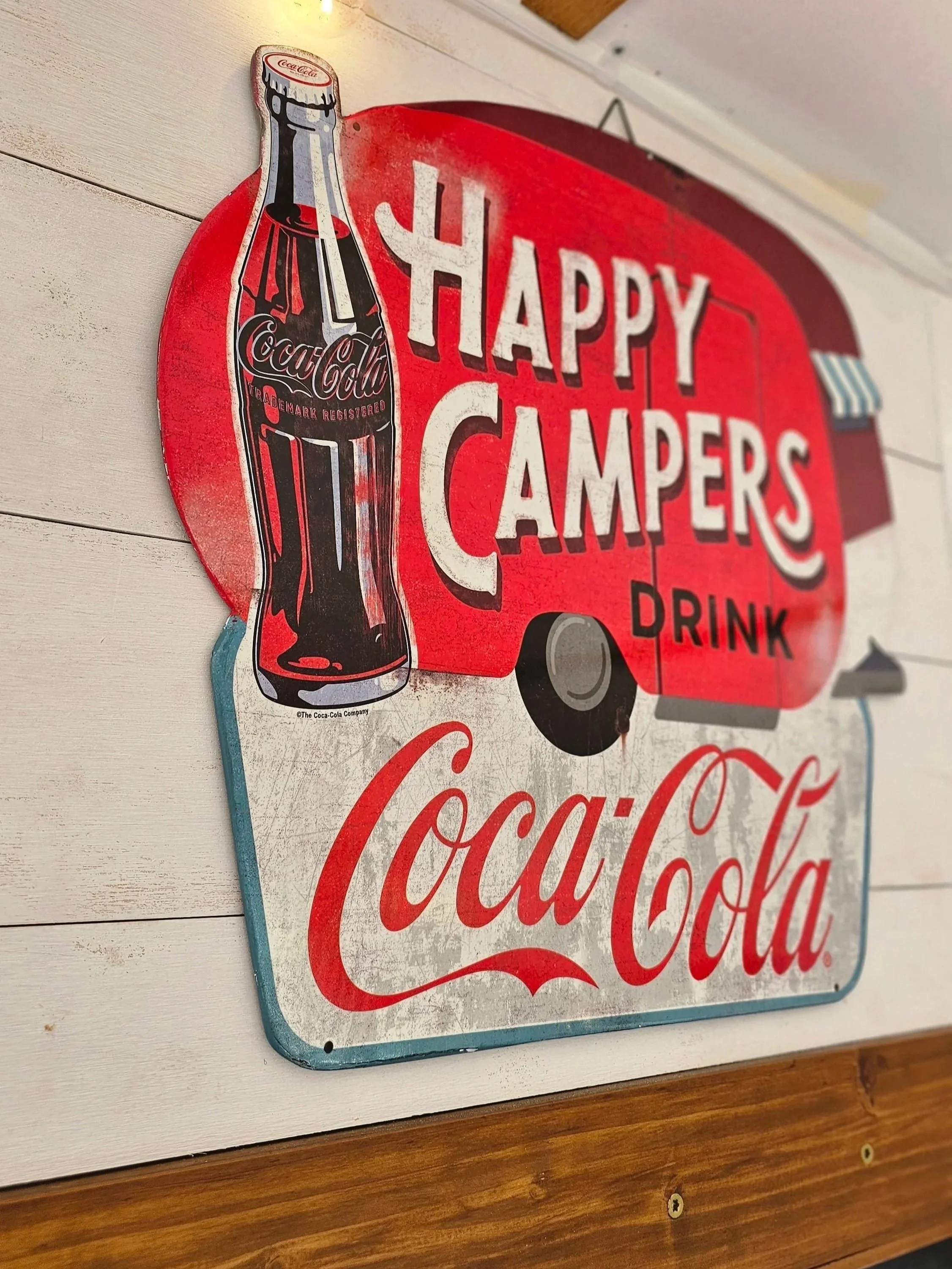 A vintage-style sign featuring a Coca-Cola bottle and the text 'HAPPY CAMPERS' and 'DRINK' on a red background, mounted on a white wall.