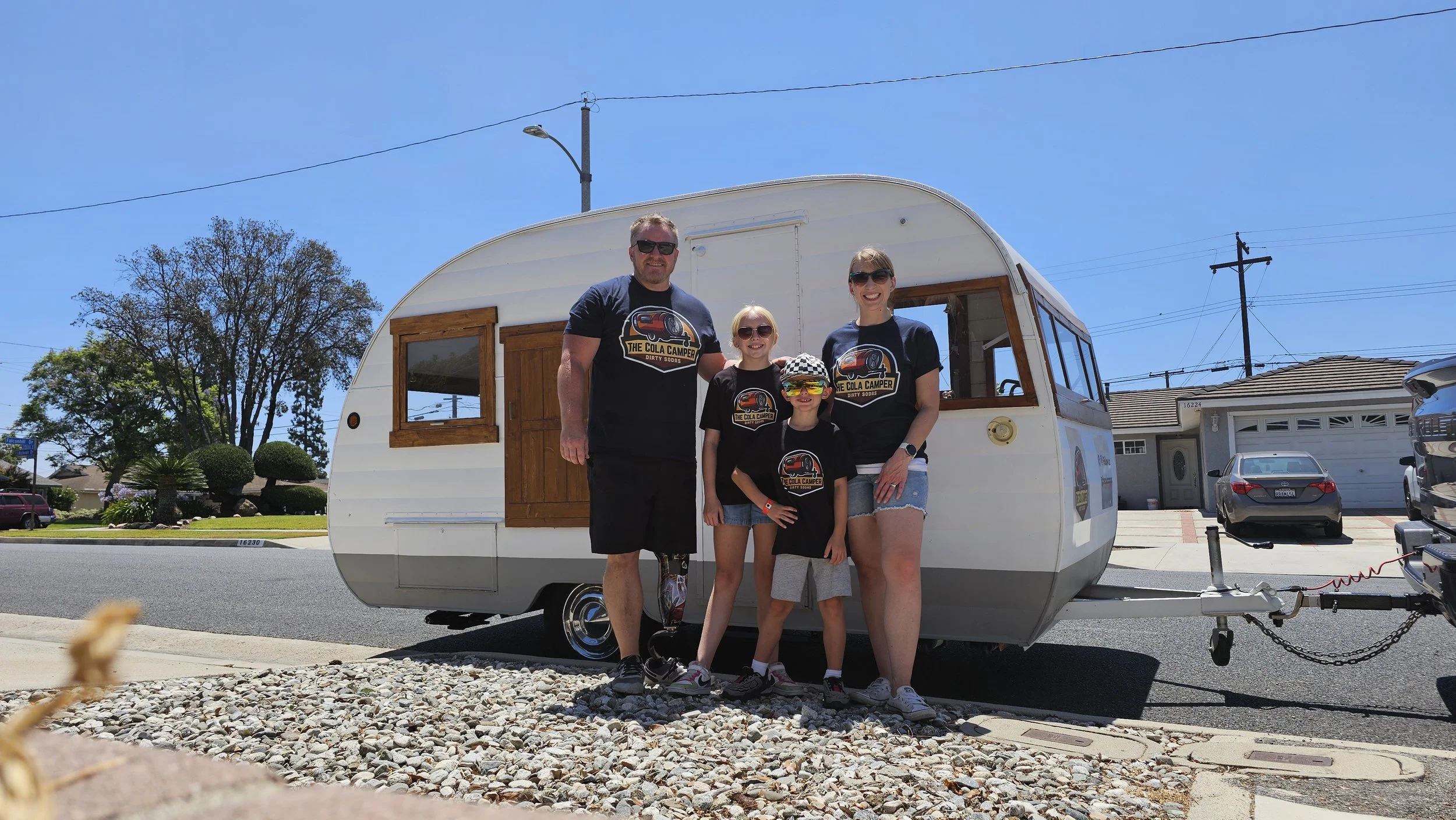 A family of four standing in front of a vintage camper trailer on a sunny day, with houses and trees in the background.