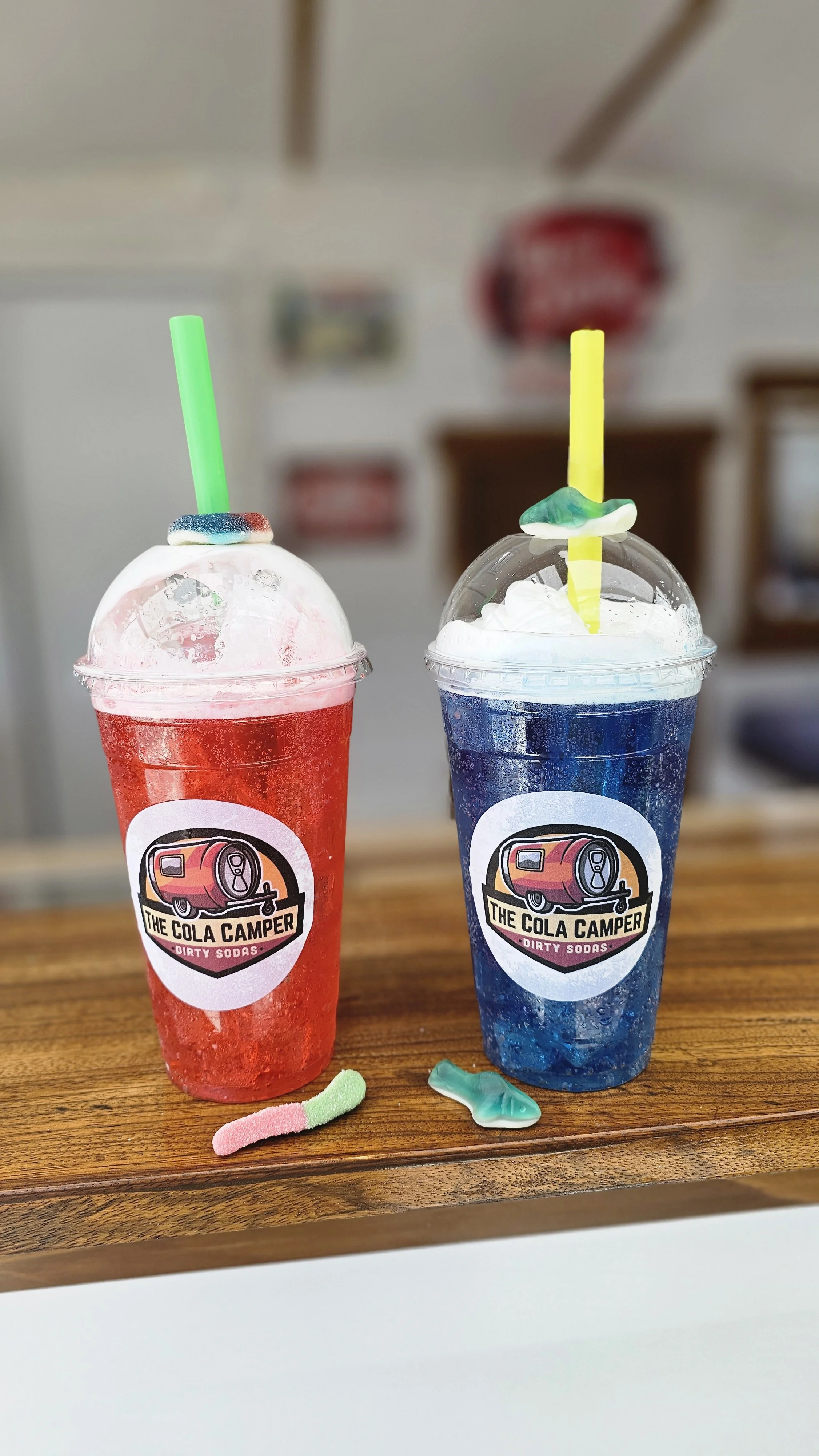 Two colorful soda drinks in plastic cups with dome lids, one red and one blue, each with a neon straw and gummy worms on top, on a wooden surface. Both cups have a logo for 'The Cola Camper - Dirty Sodas'.
