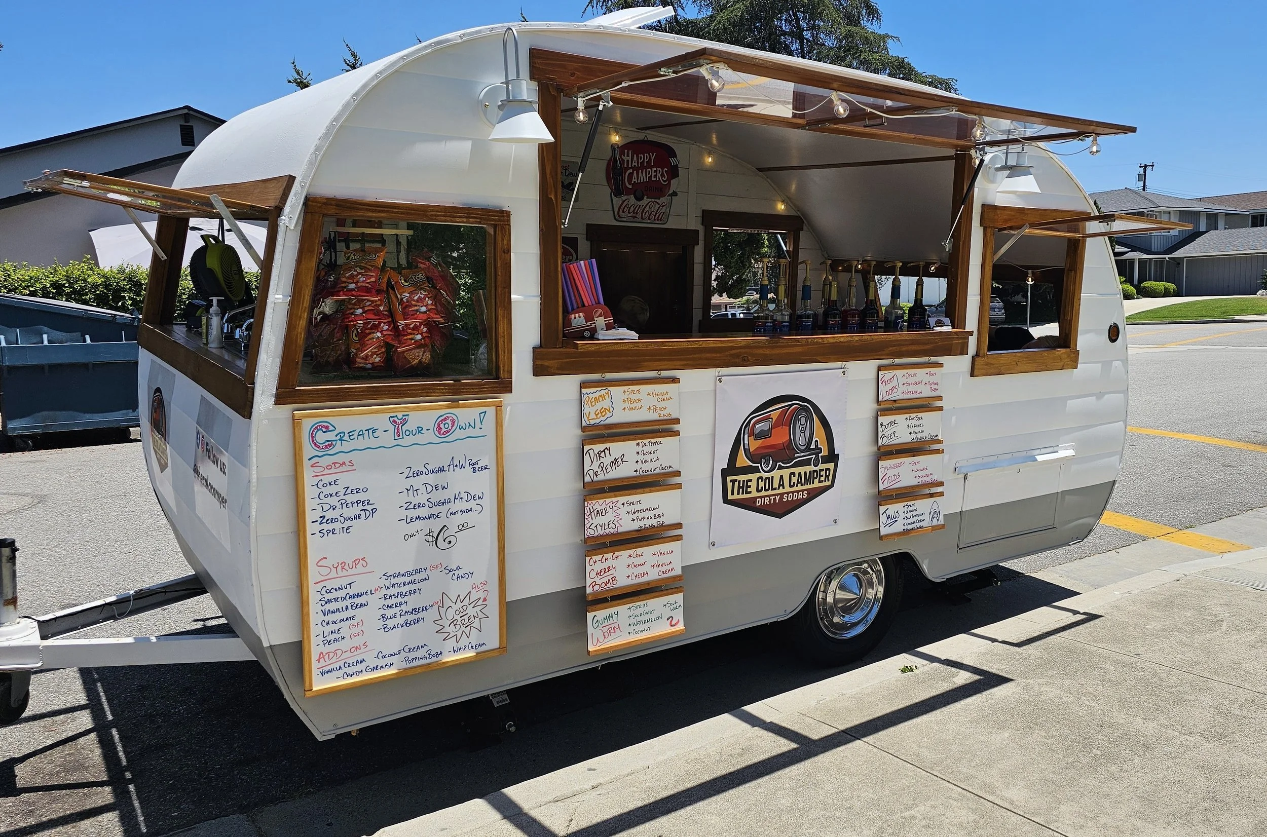A white vintage camper trailer converted into a small soda and ice cream stand named 'The Cola Camper Dirty Sodas.' The stand displays handwritten menus for various syrup flavors and toppings, with bottles of flavored syrups visible inside. The setup is outdoors on a sunny day with houses and a clear blue sky in the background.