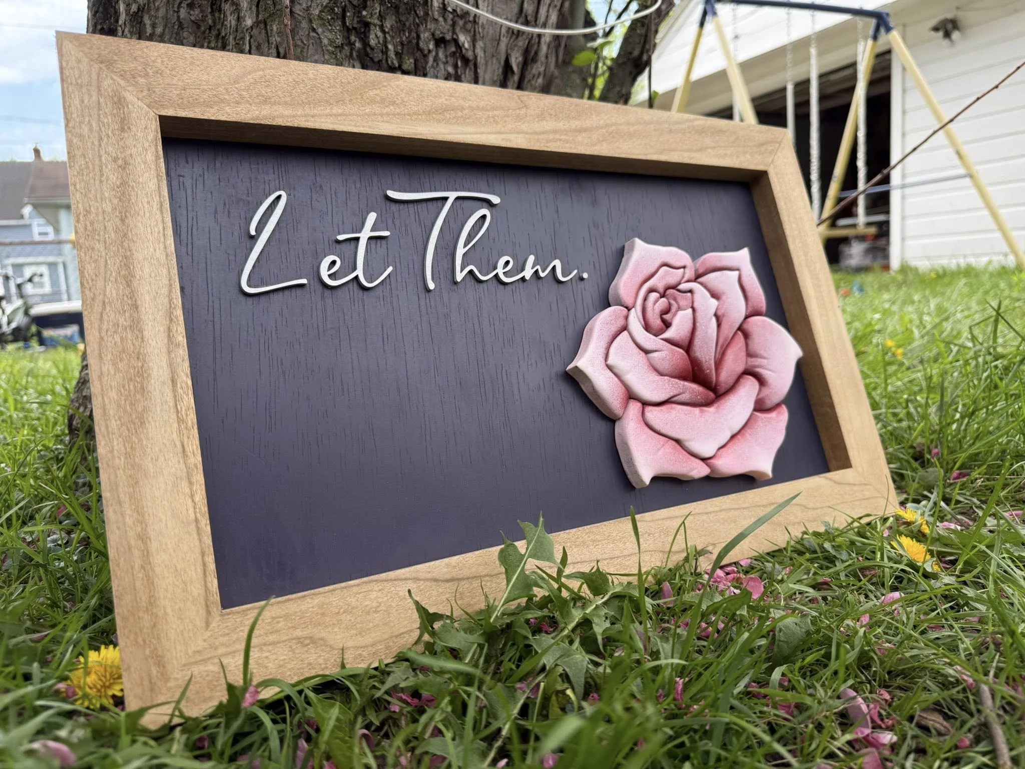 A wooden-framed blackboard with the words "Let them" in white cursive and a pink rose decoration, placed outdoors on grass with trees and a house in the background.