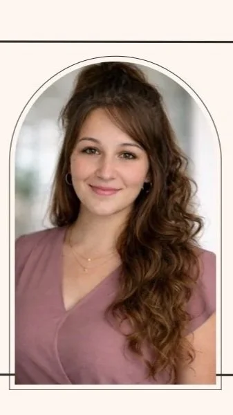 Young woman with long, wavy brown hair smiling, wearing a mauve top, standing in front of a blurred background.