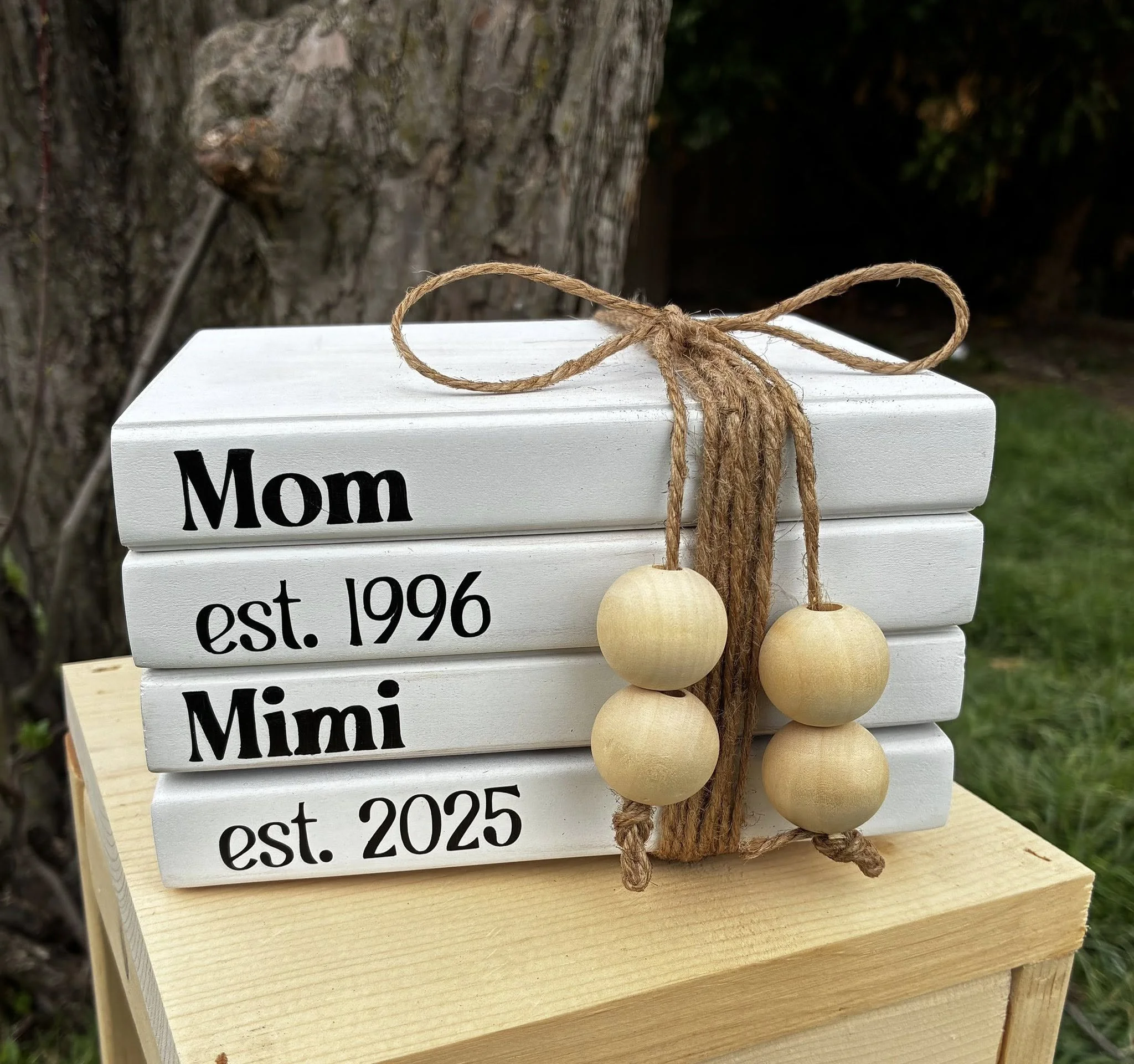 A wooden plaque labeled 'Mom' with phone number and anniversary date, decorated with a jute bow and wooden beads, placed outdoors near a tree.
