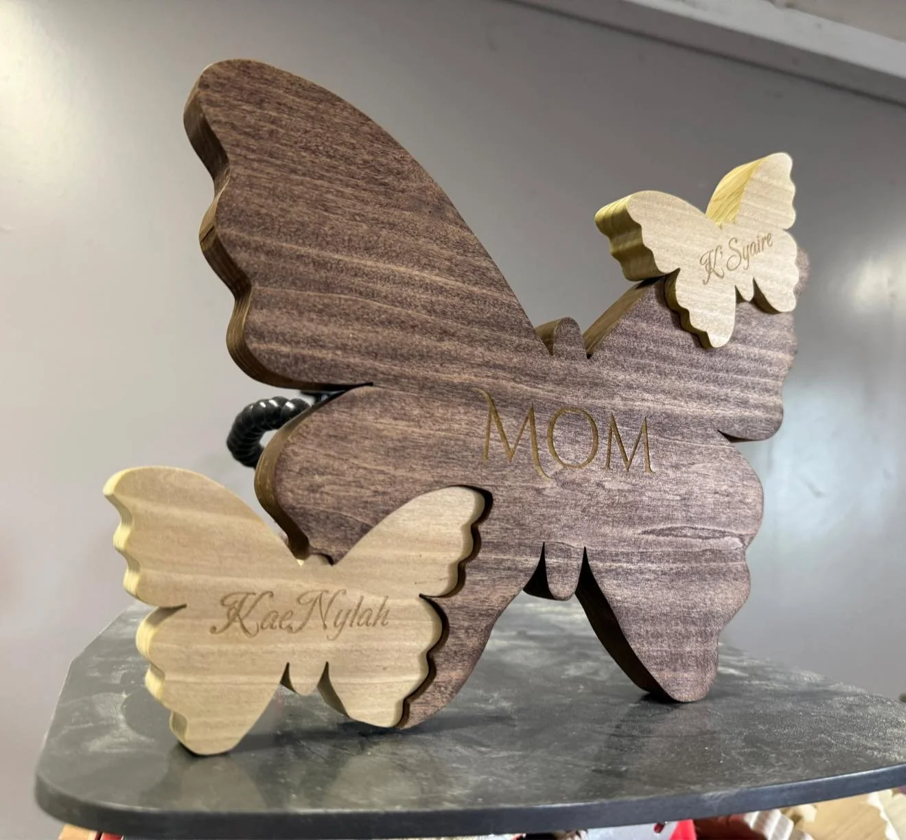 Wooden butterfly decorative piece with the word 'MOM' engraved in the center. Two smaller wooden butterflies with names 'Kaas Nylah' and 'K. Square' engraved on them, attached to the larger butterfly.