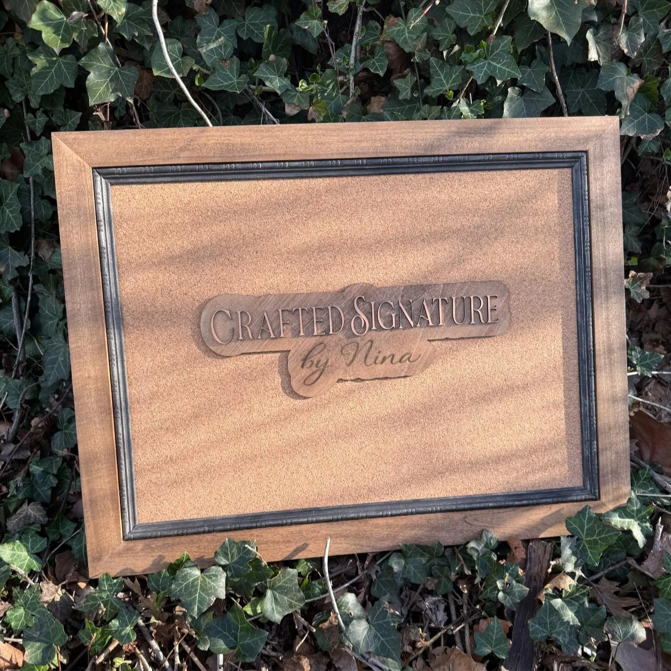 Wooden sign with engraved text reading 'Crafted Signature by Nina' placed outdoors against a backdrop of green ivy leaves.