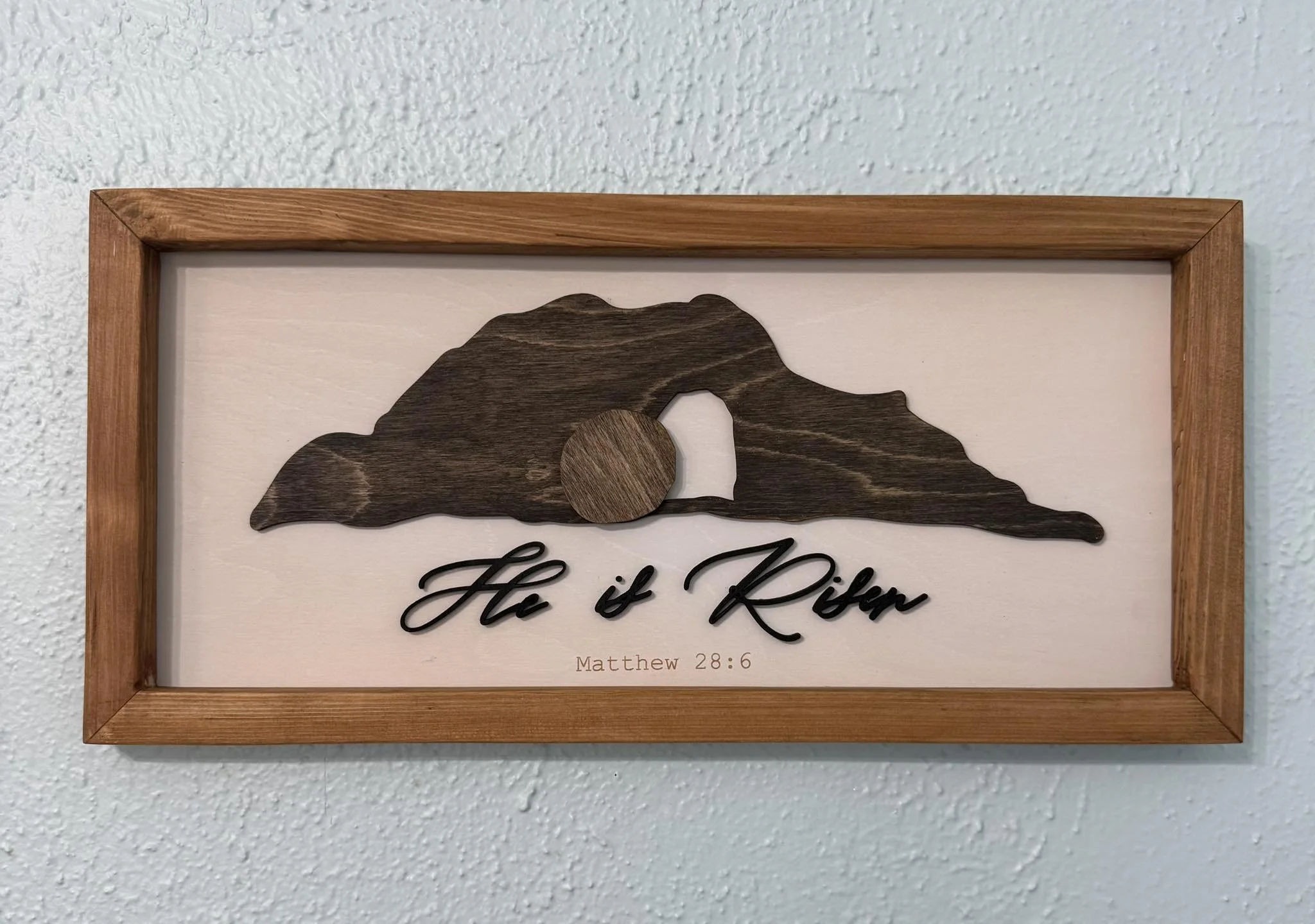 Framed artwork featuring a mountain silhouette made of wood with a circular sun and the inscription 'He is Risen' in black cursive letters, and a Bible verse reference Matthew 28:6 at the bottom.