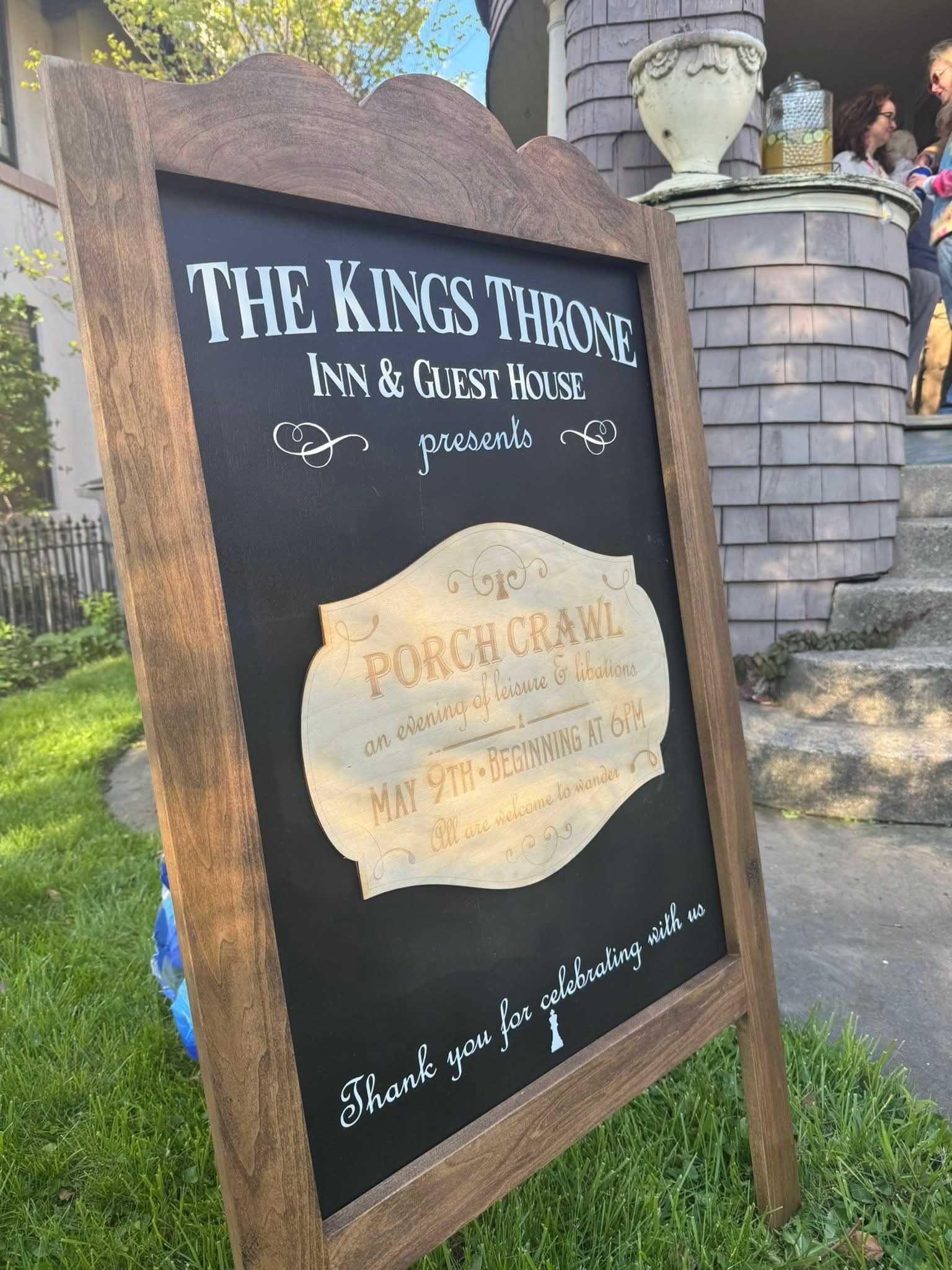 A chalkboard sign for The Kings Thorne Inn & Guest House with a decorative wooden frame. The sign has a note of thanks and details about an event, with a background of a house porch and people standing at a table, and a large urn and a jar with lemon