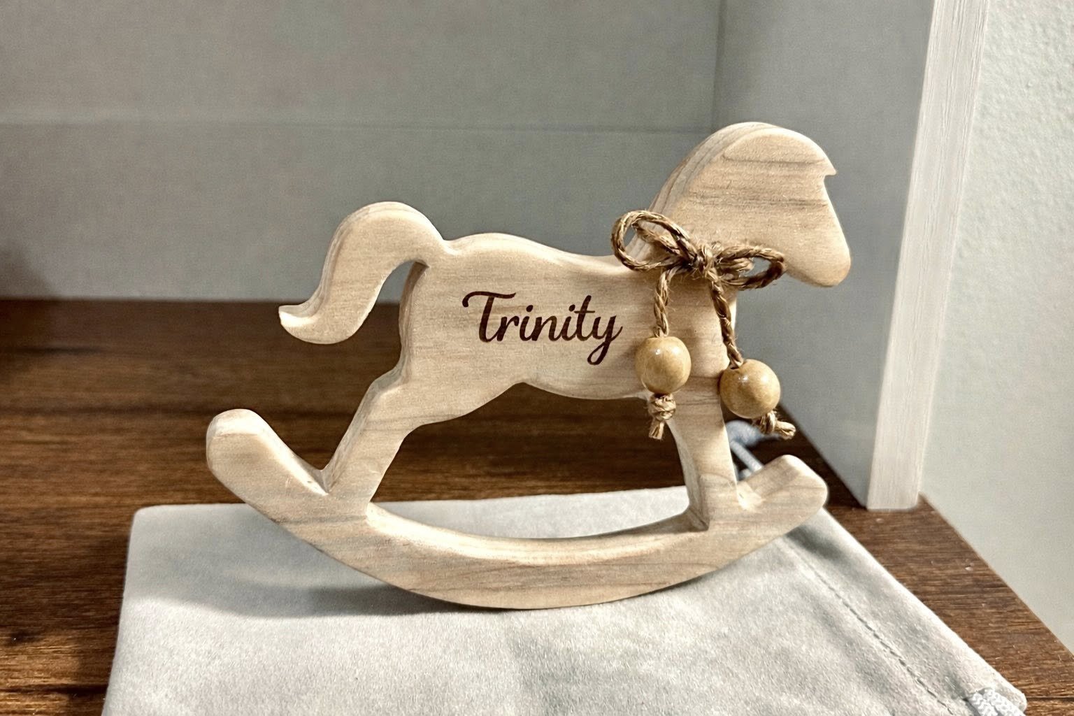 Wooden rocking horse ornament with the word 'Trinity' engraved, decorated with a bow and wooden beads, placed on a surface.