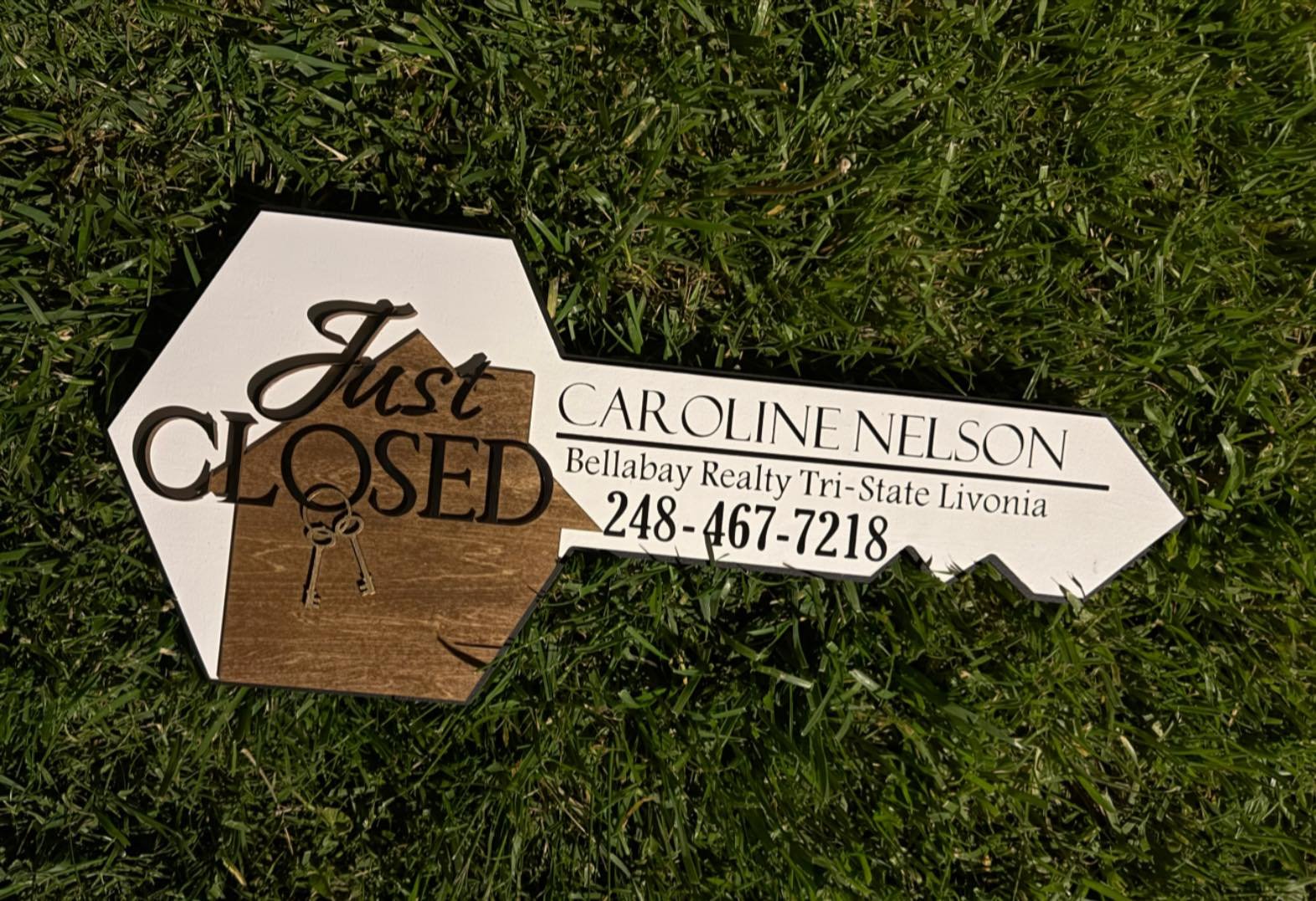 A yard sign indicating a property is 'Just Closed' with the realtor's contact information, Caroline Nelson, from Bellabay Realty Tri-State Livonia, and a phone number 248-467-7218, lying on grass.
