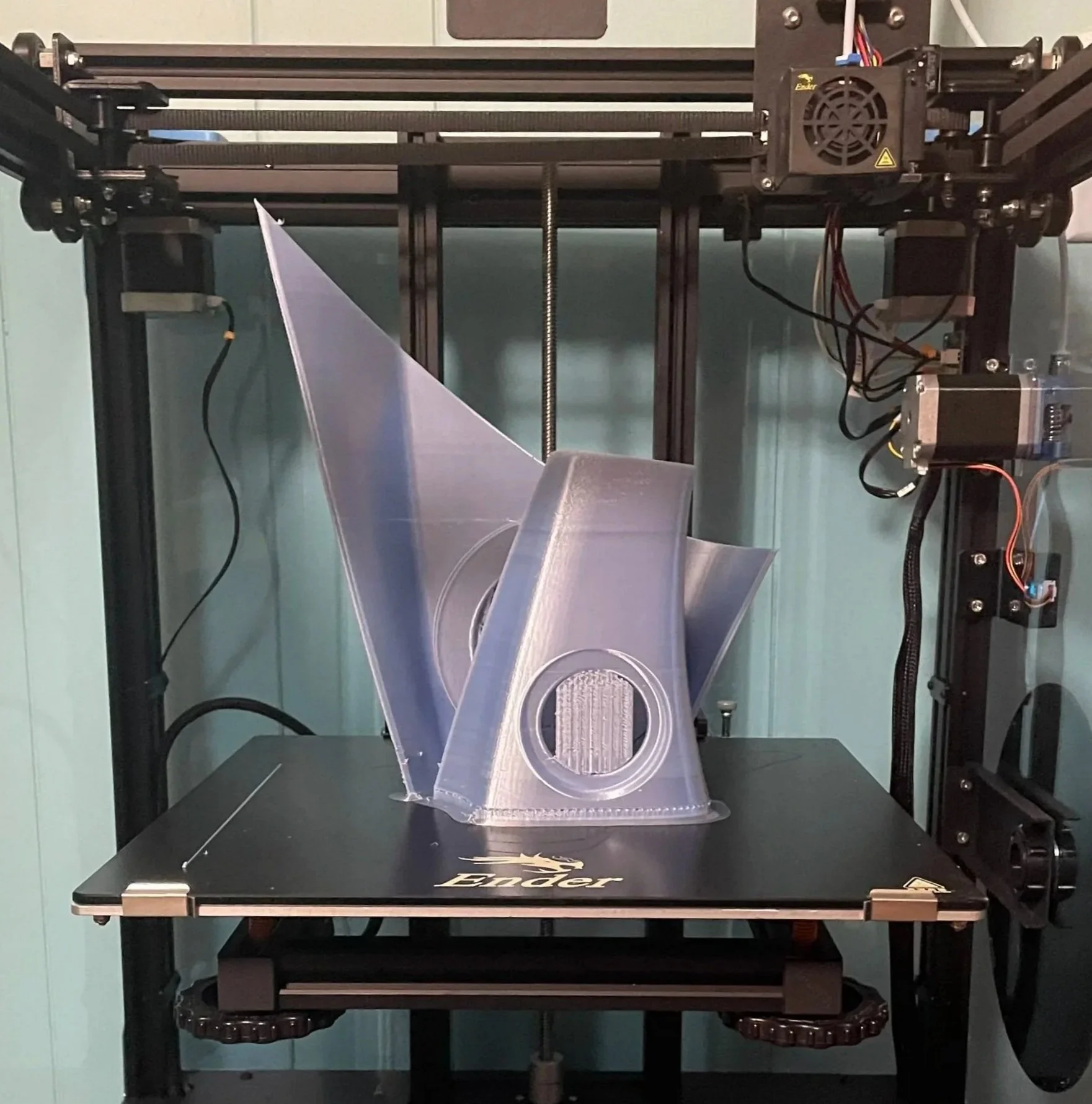 A 3D printer with a printing bed and an extruder nozzle. The printer is branded 'Ender' with a dragon logo, and the setup includes various wires and mechanical components.