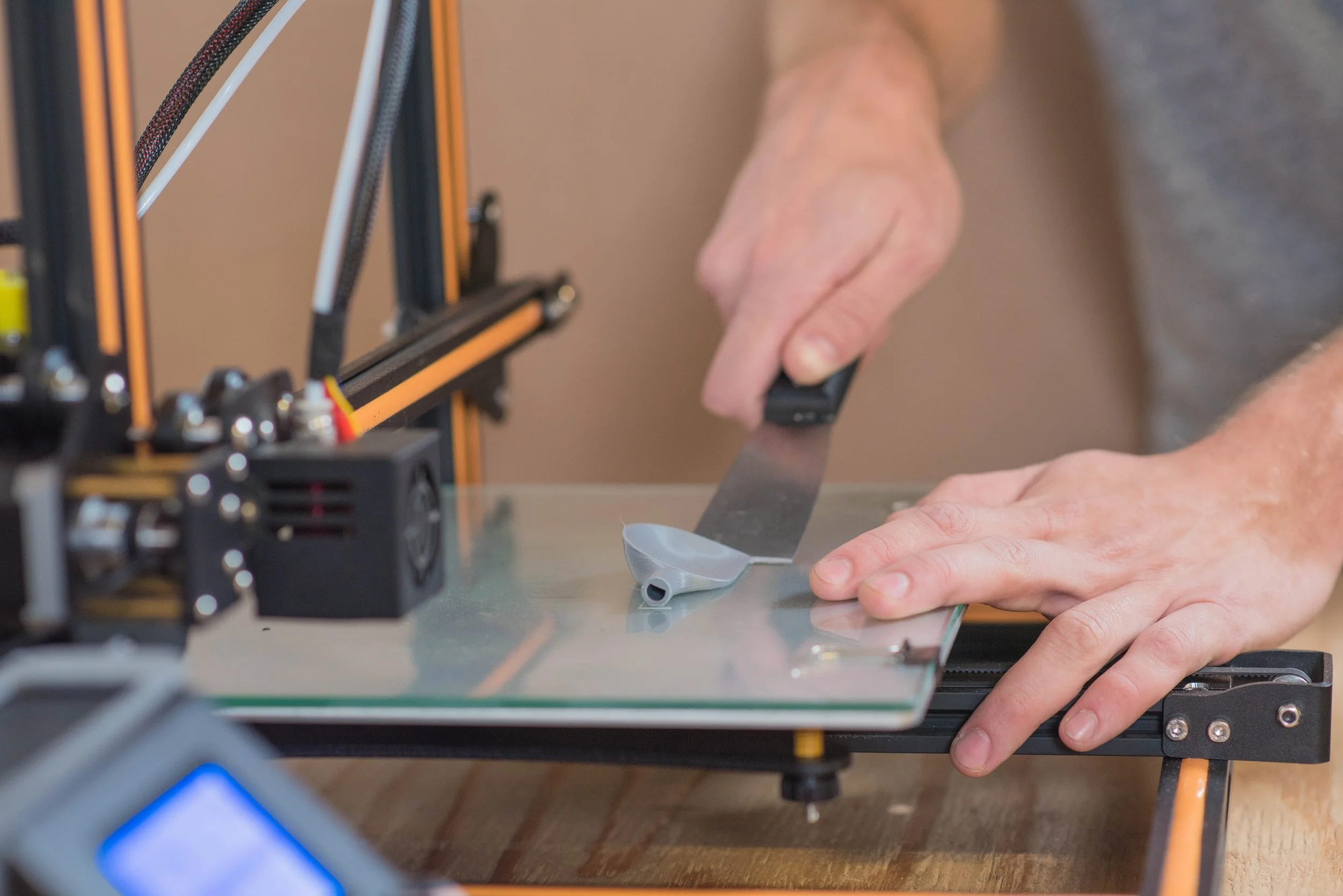 A person working on a 3D printer, adjusting or trimming a part with a small tool, with the printer's components and frame visible in the background.