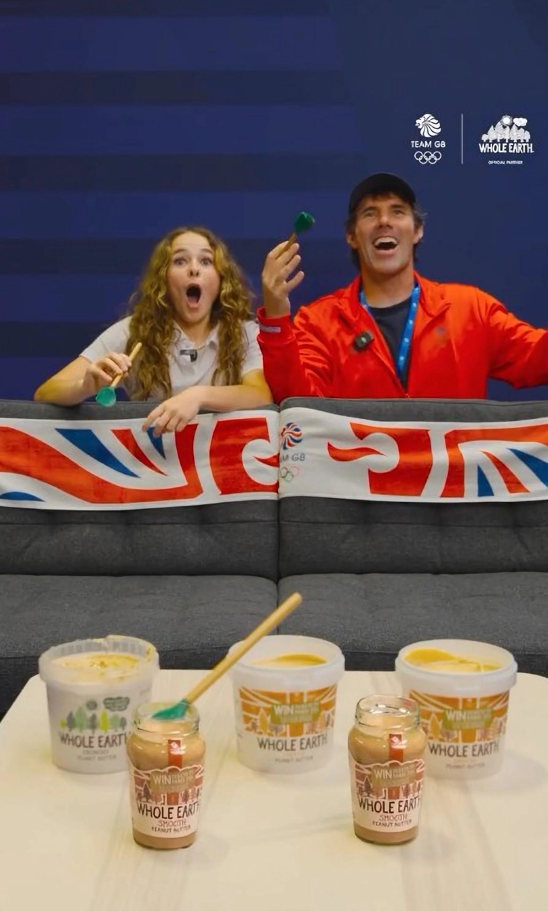 Two people, a young woman and an older man, sitting at a table with various containers of Whole Earth yogurt in front of them. The young woman appears surprised, holding a spoon, while the man is laughing. The background has a blue wall with logos of Team GB and Whole Earth.