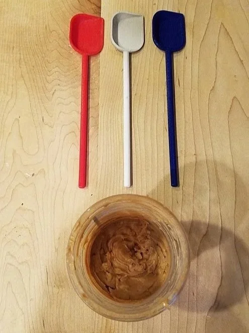 Three plastic spatulas in red, white, and blue are placed on a wooden surface above a clear jar containing peanut butter