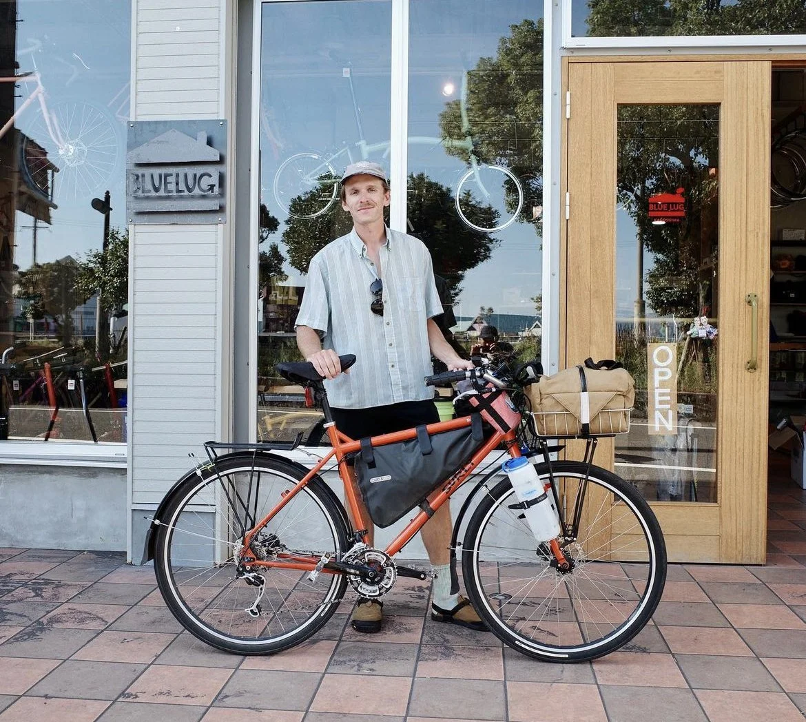 A man standing with a bike outside a storefront, wearing a light-colored shirt, shorts, a cap, and sandals, with a water bottle attached to the bike. The store window behind has bicycles and an 'OPEN' sign on the door. Blue Lug Japan