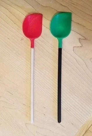 Two spoon-shaped objects, one red with a white handle and one green with a black handle, placed on a wooden surface.