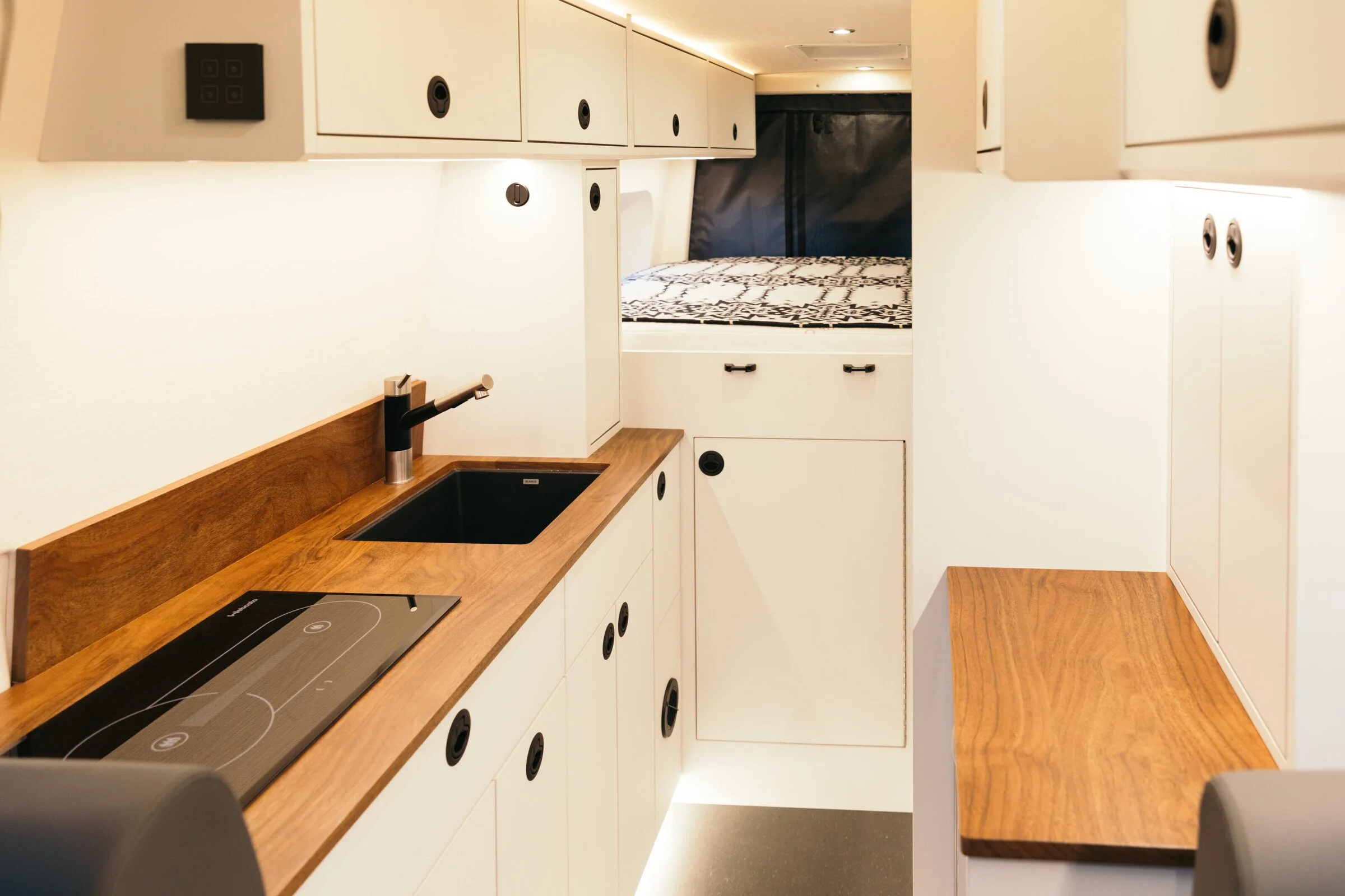 A compact camper kitchen with white cabinets, a black sink with a chrome faucet, a black electric stovetop, and a wooden countertop, leading to a sleeping area with a bed and black privacy curtain.
