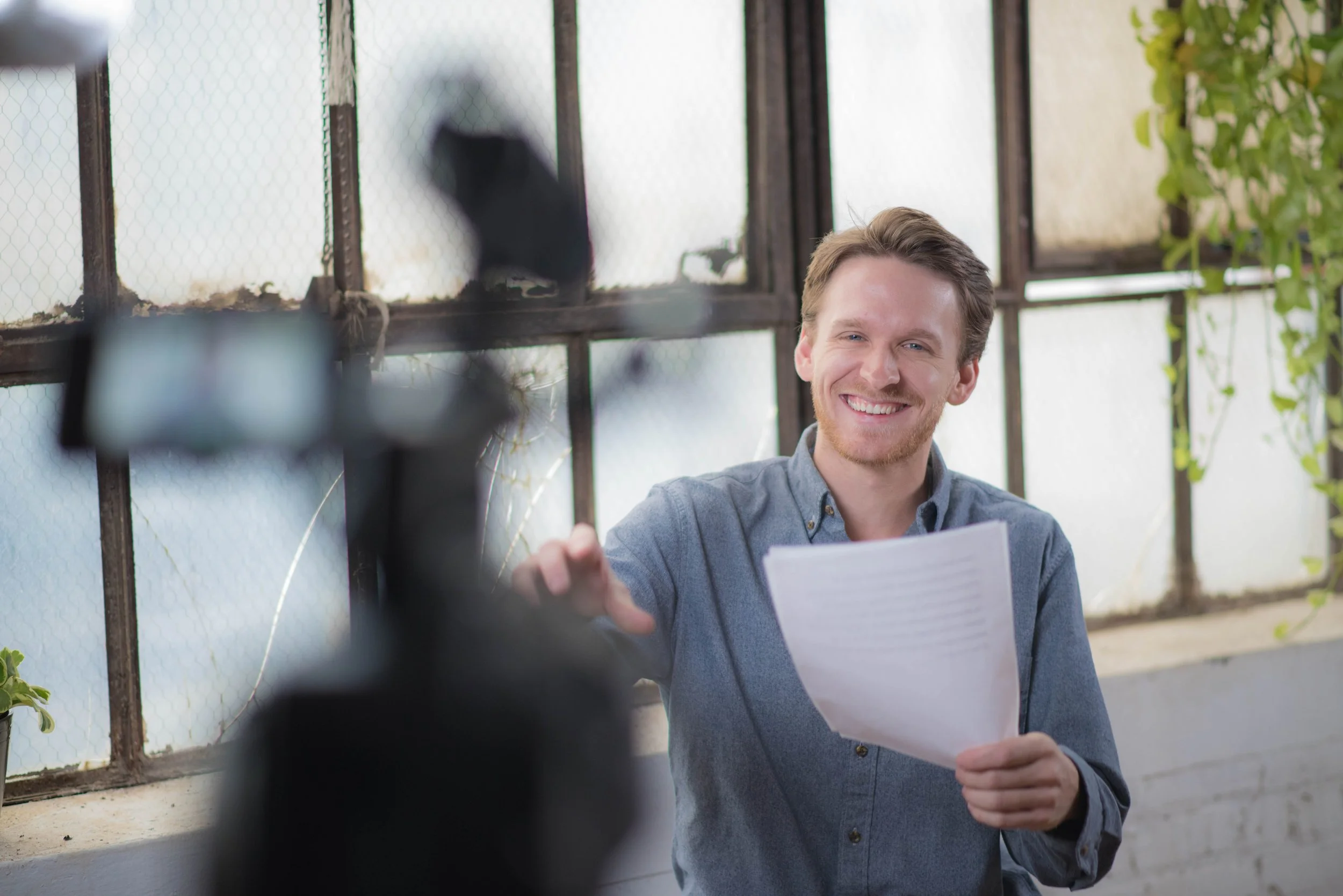 A young man with short light brown hair and a beard smiling while taking a selfie inside a greenhouse, with plants and large windows in the background.