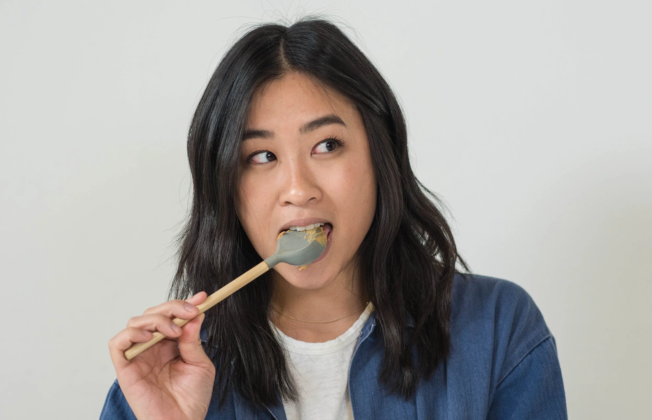A young woman with long black hair and a blue denim jacket is eating food with a wooden spoon.