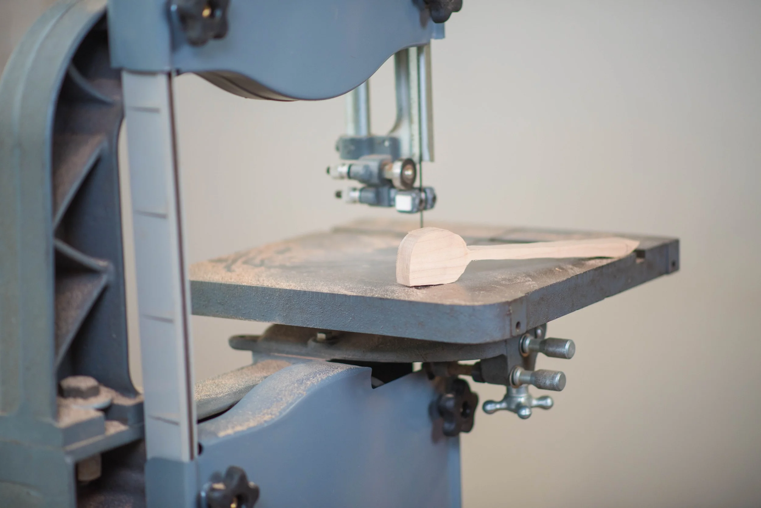 A bandsaw cutting a piece of light-colored wood on a gray worktable.