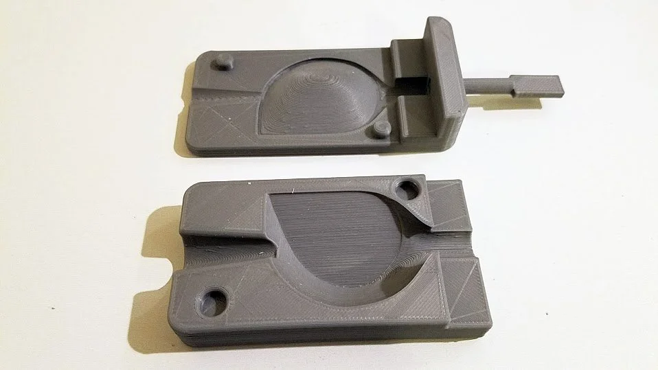 Two gray 3D-printed parts with semicircular recesses and mounting holes, one with a sliding component attached.