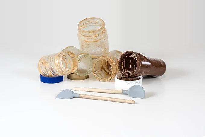 Empty glass jars, one with chocolate spread, and two small spatulas on a white background.