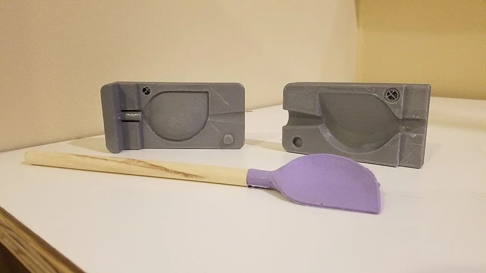 Two gray plastic objects resembling molds or clips and a wooden stick with a purple piece of modeling clay attached.
