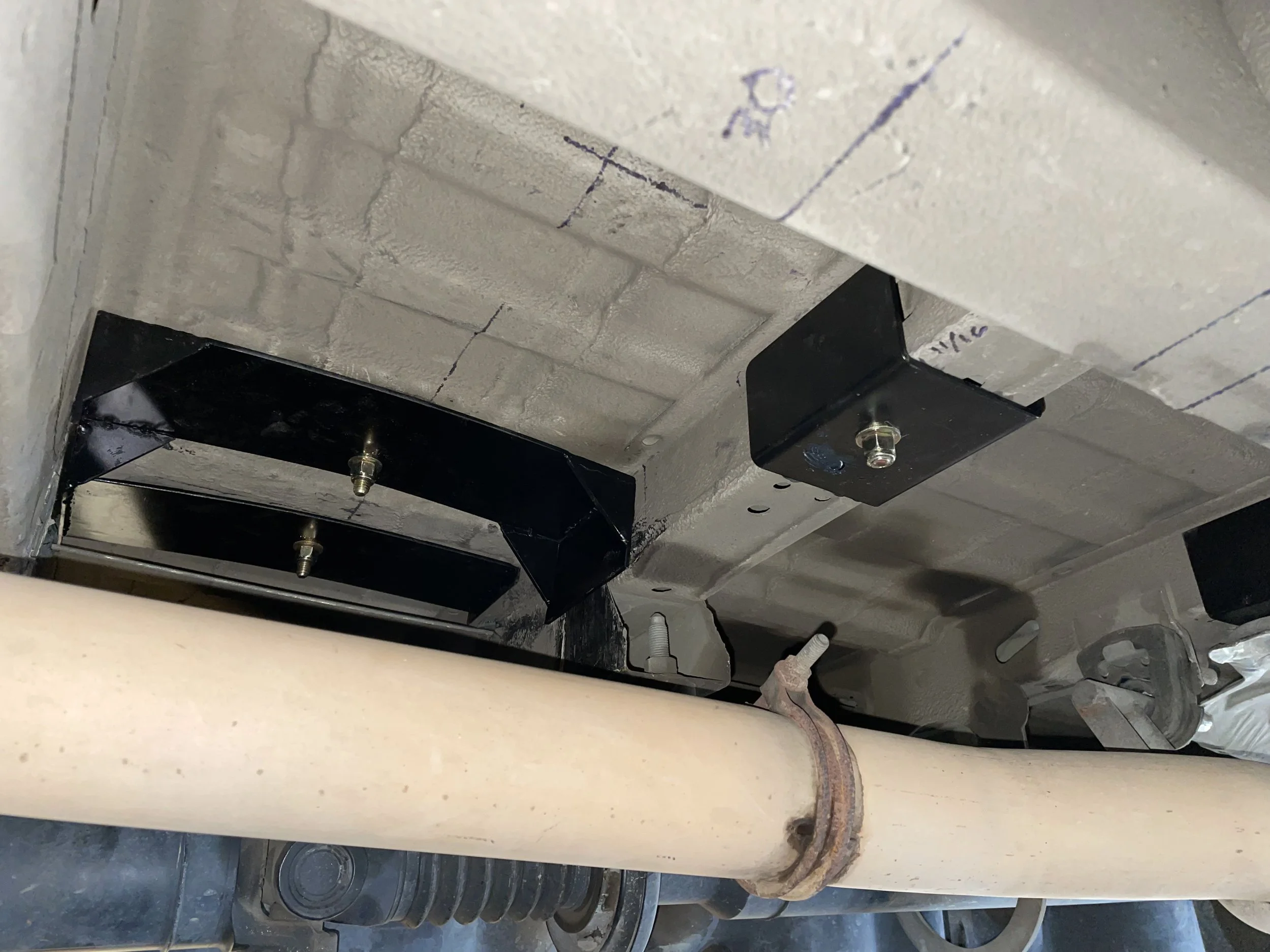 Underbelly of a vehicle showing a large metal exhaust pipe (or similar component), some black brackets, and part of the vehicle's frame with bolts and metal parts.