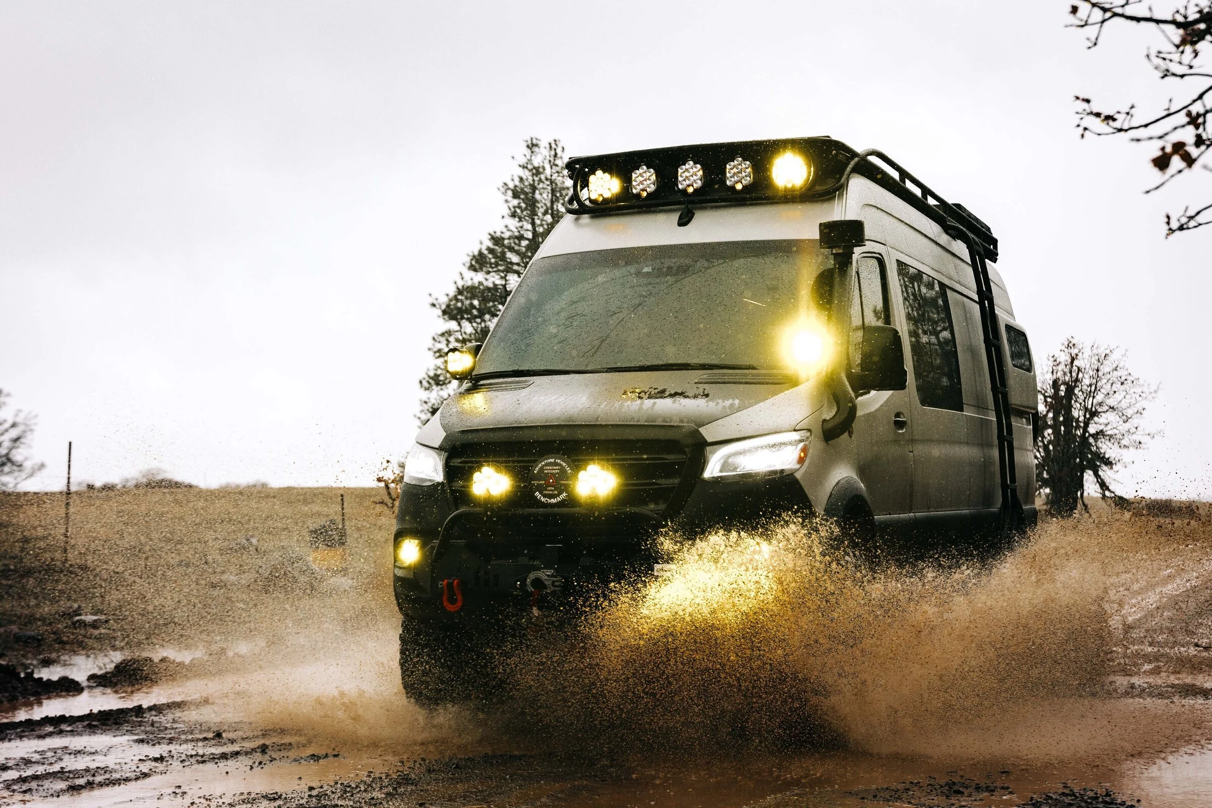 A gray off-road vehicle driving through water, with its lights on during overcast weather, trees in the background.