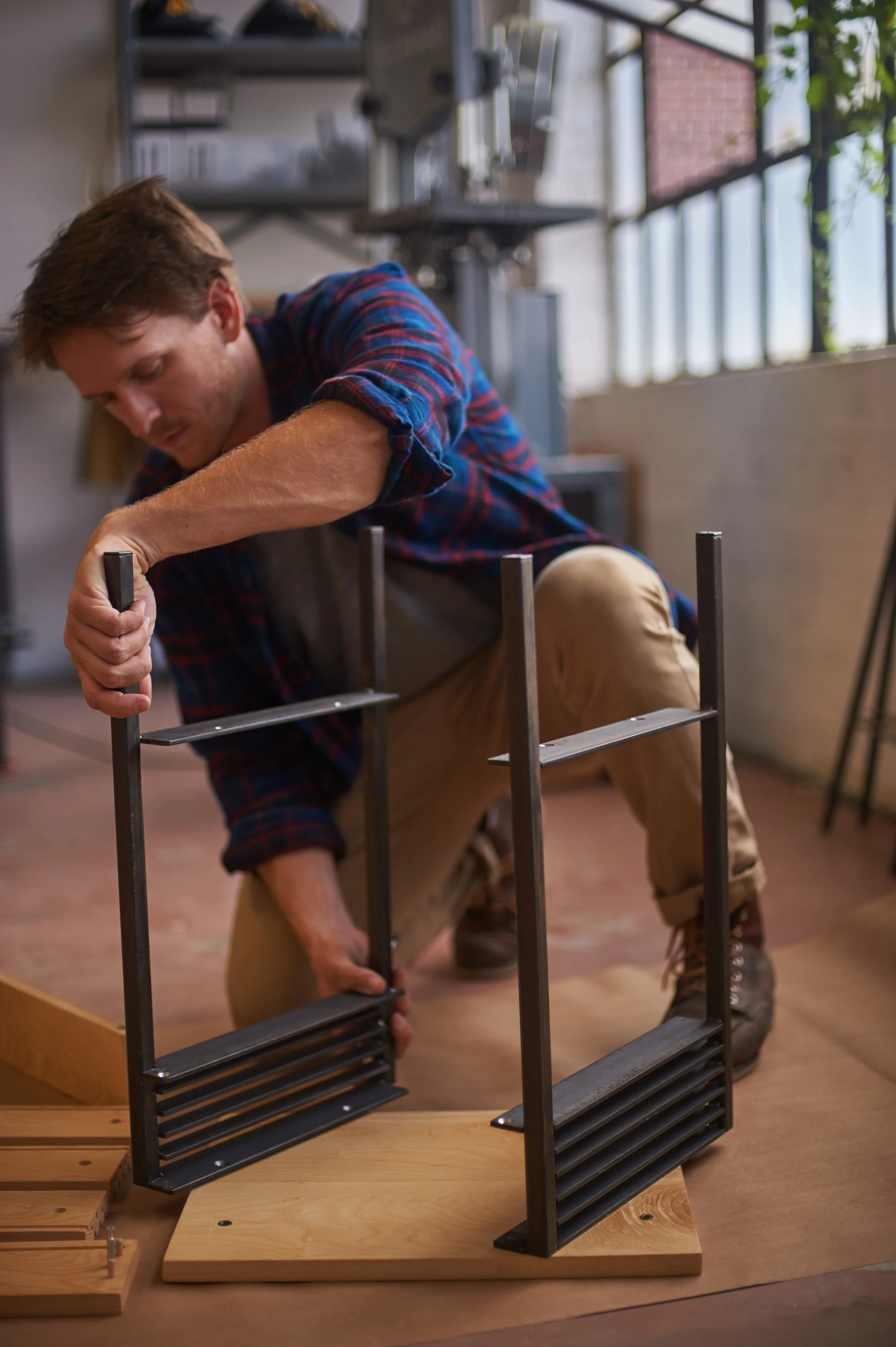 Man working on a woodworking project in a workshop with large windows, assembling a structure with metal and wood components.