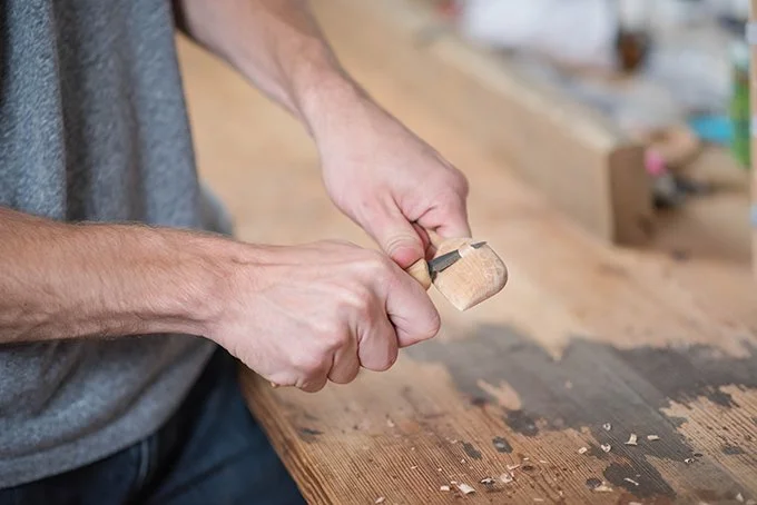 A person holding a small hammer and working on a piece of wood on a workbench in a woodworking shop.