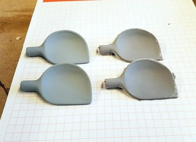 Four gray plastic objects that look like small spatulas or scoops, with flat, rounded ends and short handles, placed on a grid-lined sheet of paper.