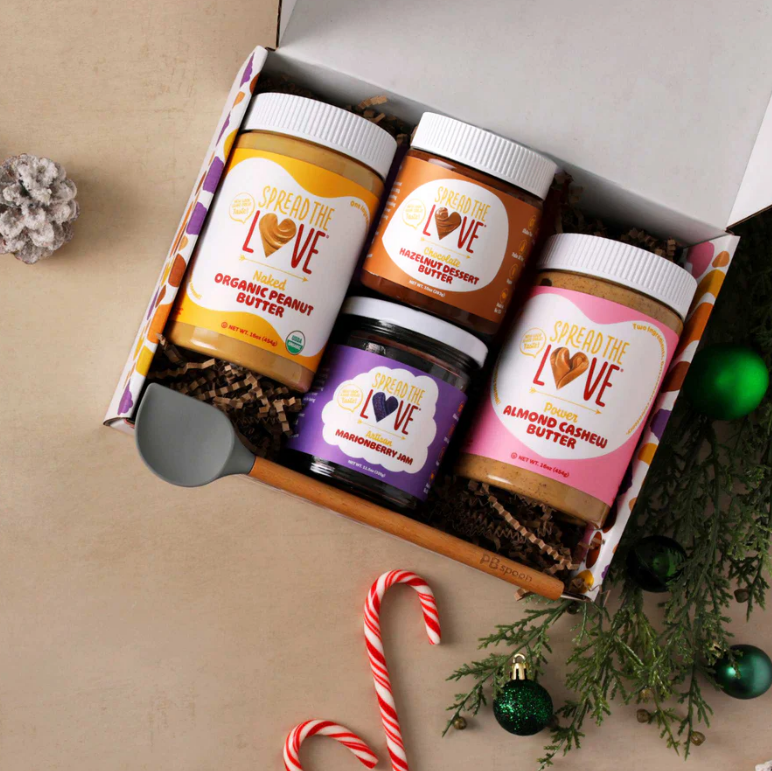 A gift box containing four jars of various spread butters, including organic peanut butter, hazelnut dessert butter, and almond cashew butter, surrounded by holiday decorations like a candy cane, pine branches, and ornaments.